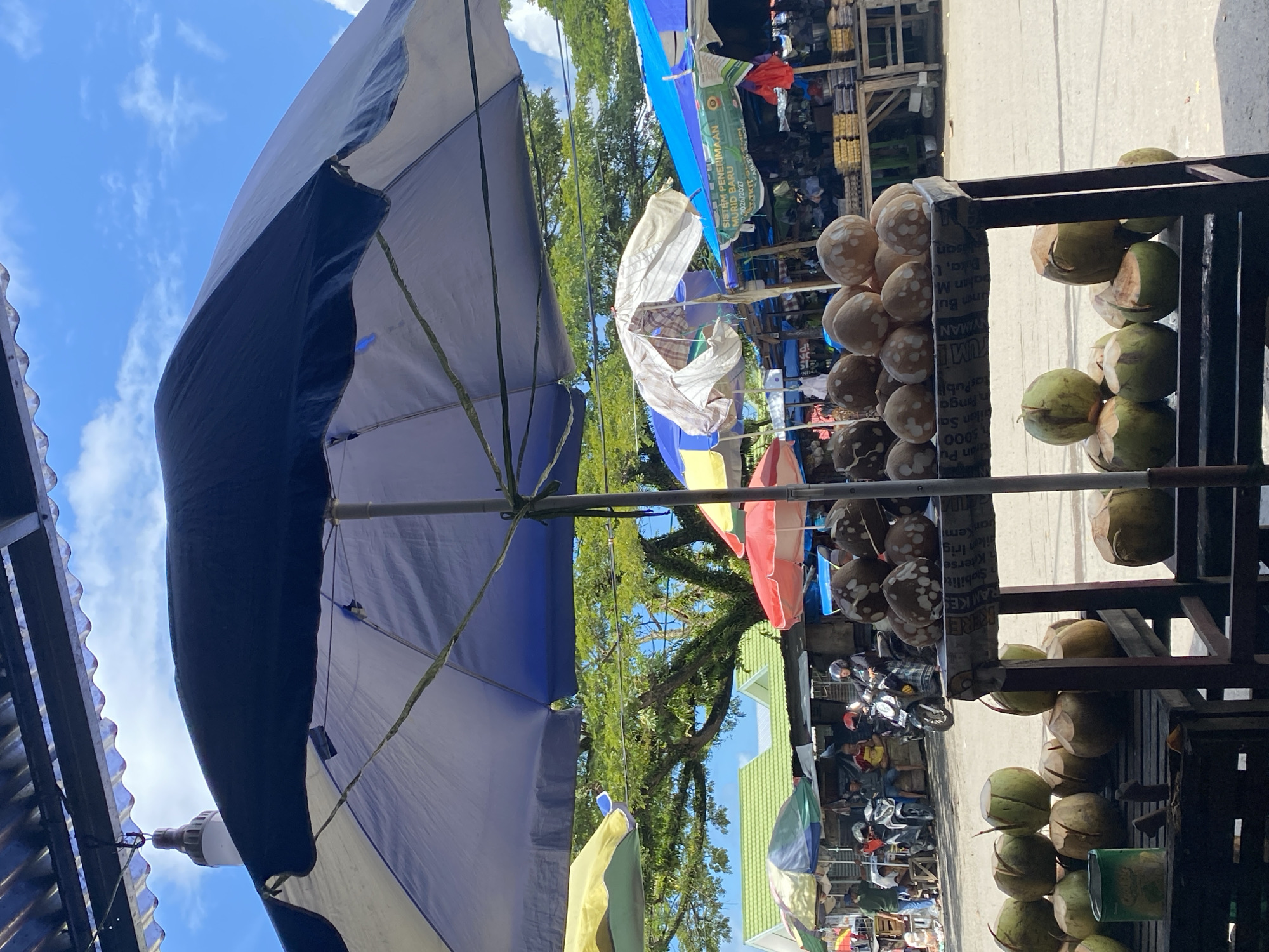 An outdoor market scene in Ta', Indonesia, is depicted under a bright, cloudy sky. In the foreground, a stall displays a large quantity of coconuts, some whole and some cut open. A dark blue and white umbrella shades the stall. Beyond this, a series of market stalls and temporary structures are visible, covered by various colored umbrellas, including yellow, red, and blue. People are present, some tending to the stalls, others on motorcycles, suggesting active commerce. In the background, a large, leafy tree provides shade, and some buildings with green roofs are discernible. The overall atmosphere is that of a bustling local marketplace.