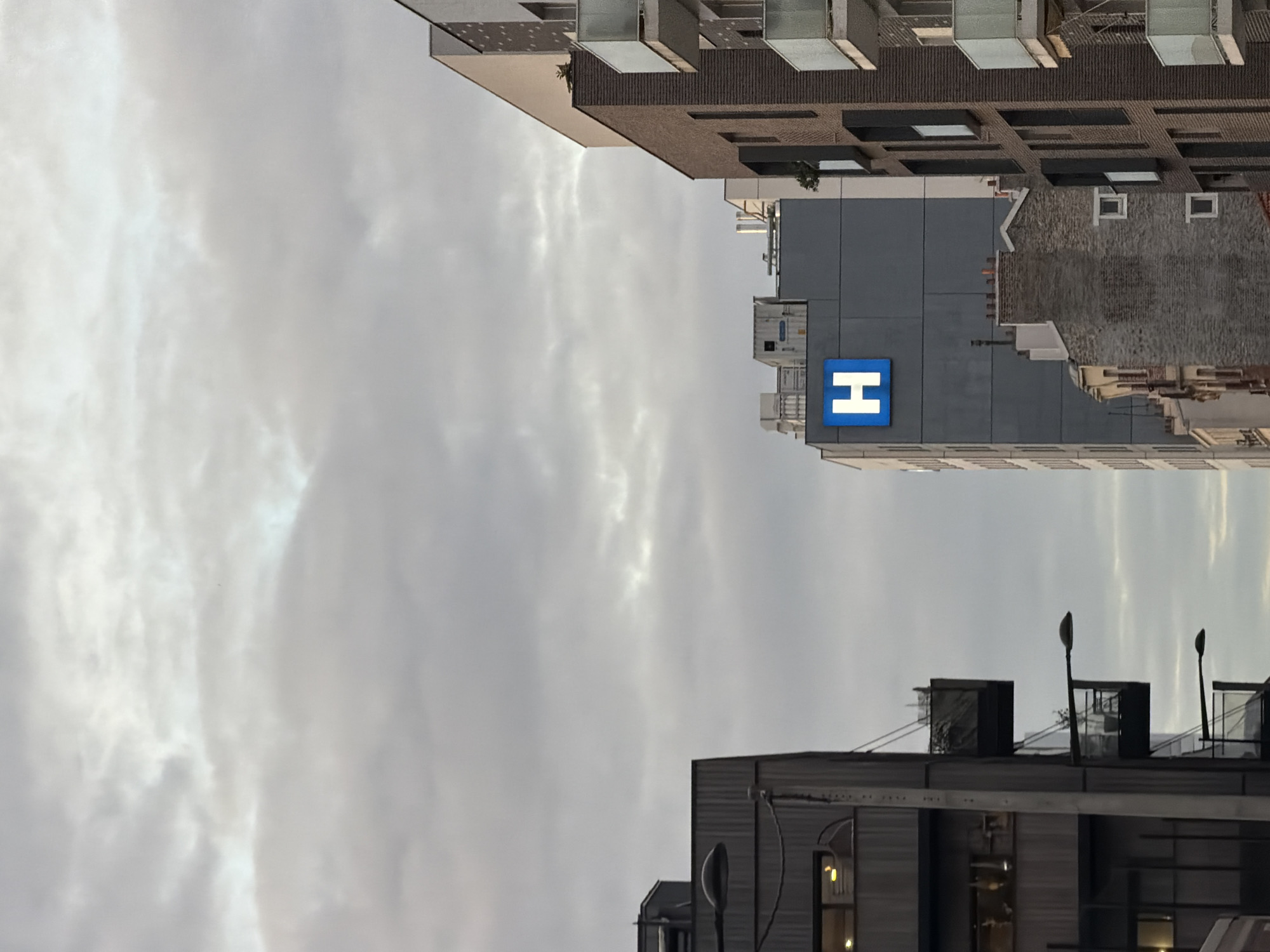 The image depicts a view of the sky above Aubervilliers, France, framed by buildings. In the foreground, the upper portion of a grey building with horizontal lines is visible, punctuated by windows and rooftop structures, including streetlights and what appears to be mechanical equipment. Above and slightly to the right, a taller, grey building has a large blue square with a white "H" on it, likely indicating a hospital or medical facility. Above this, the upper portions of several other buildings of varying heights and architectural styles are visible, some with balconies and others with what appears to be brick facade. The sky is overcast with heavy cloud cover, giving the scene a subdued, neutral color palette.