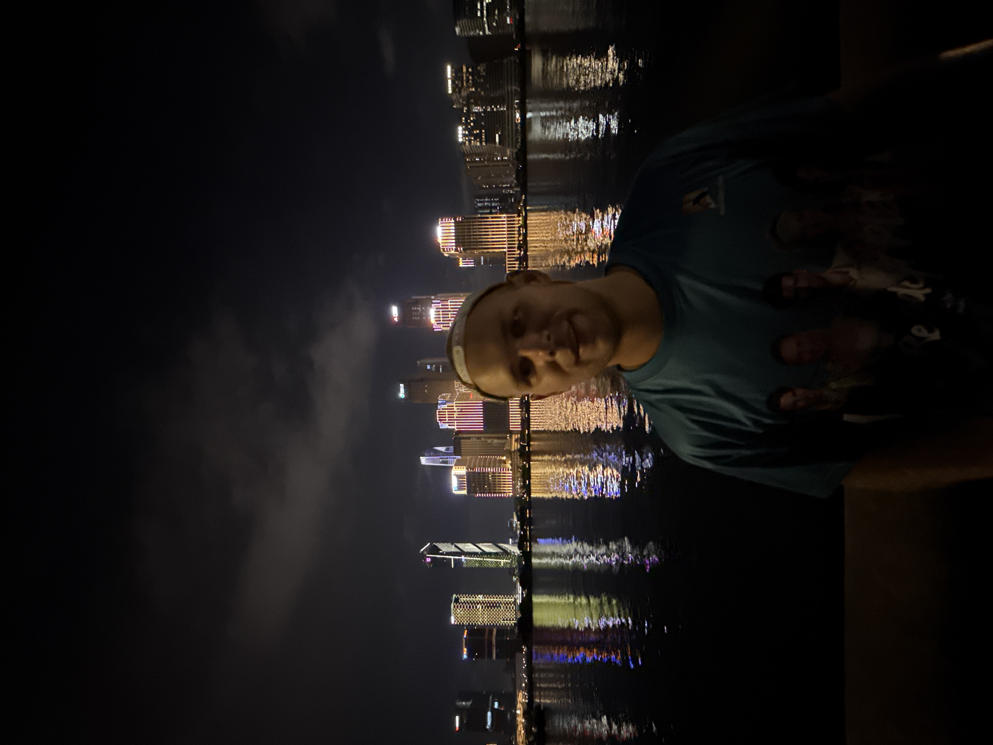 The image displays a nighttime scene in Haizhu District, Guangzhou, China, featuring a Caucasian male standing on the south bank of the Pearl River, looking north towards the illuminated cityscape of Tianhe District. The man, appearing to be in his 20s or 30s, is positioned slightly off-center to the right, facing the viewer with a subtle smile. He wears a light blue crew-neck t-shirt with faded, indistinct images of multiple individuals printed on the front, and a white headband.
Behind the man, the Pearl River is visible, reflecting the colorful artificial lights of numerous skyscrapers that dominate the opposite bank. The buildings exhibit diverse architectural styles and are brightly lit with various patterns of yellow, red, blue, and white lights. A prominent, tall skyscraper with a distinctive illuminated zigzag or spiral design is visible towards the center-left of the skyline. The dark night sky above the city shows faint cloud formations. The reflections of the city lights create shimmering, elongated streaks on the dark water surface. The lower portion of the image reveals a dark, indistinct bank or walkway where the man is standing.