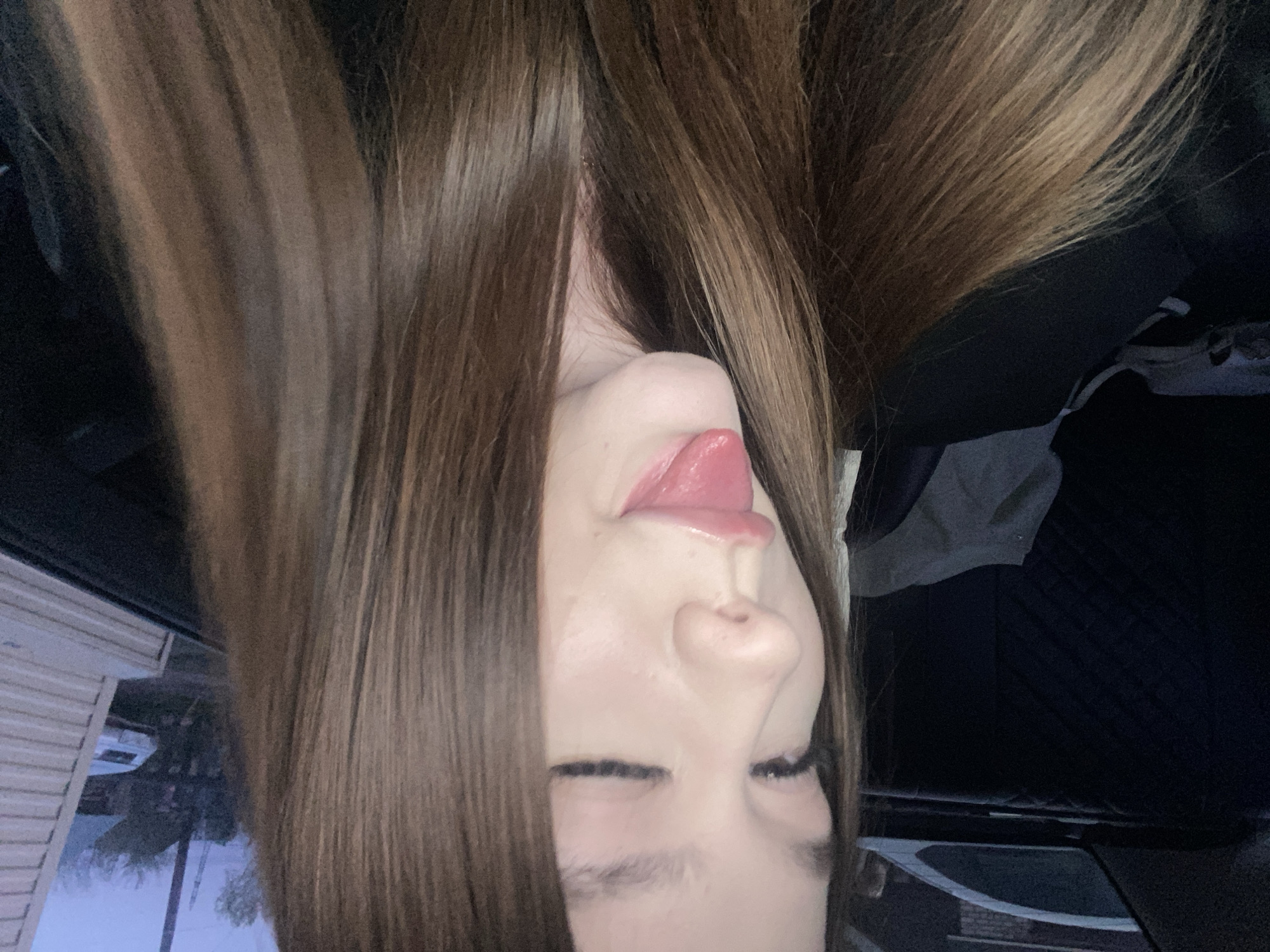 The image shows a person with long, brown hair, fair skin, closed eyes, and a tongue sticking out. The background is blurry but appears to show a window, a light-colored building, and what might be the interior of a car. The location cannot be determined as Chaikovsky, Russia.