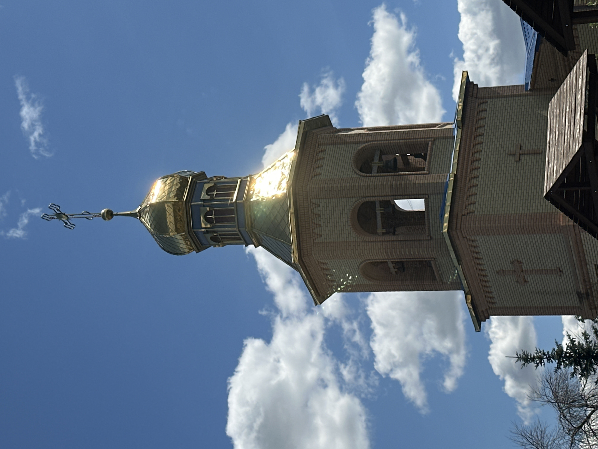 The image shows the bell tower of a church with a golden dome and a cross on top. The bell tower is made of light brown brick and has arched openings where bells are visible. The sky is blue with white clouds. The church is located in the city of Zhovte, Ukraine.