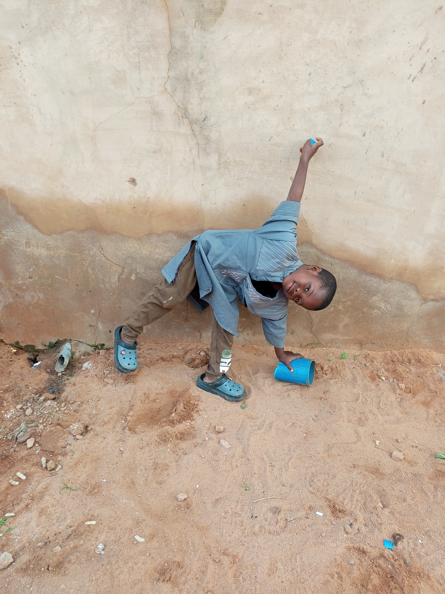 The image shows a young boy in a blue-gray tunic and brown pants with a checkered pattern, wearing black clogs, in a sandy area against a textured, light-colored wall. He is in a dynamic pose, leaning to his right with his left arm extended upwards and his right hand holding a blue cylindrical object. The wall has cracks and stains, and the ground is sandy with some scattered rocks and debris. A piece of broken pipe is visible on the left side of the image near the wall. The scene appears to be outdoors in a casual, possibly rural, setting. The boy is looking towards the camera with a slight smile. The presence of a wall suggests it might be the side of a building. Without additional context, it's impossible to definitively place this scene in Tempure, Nigeria.