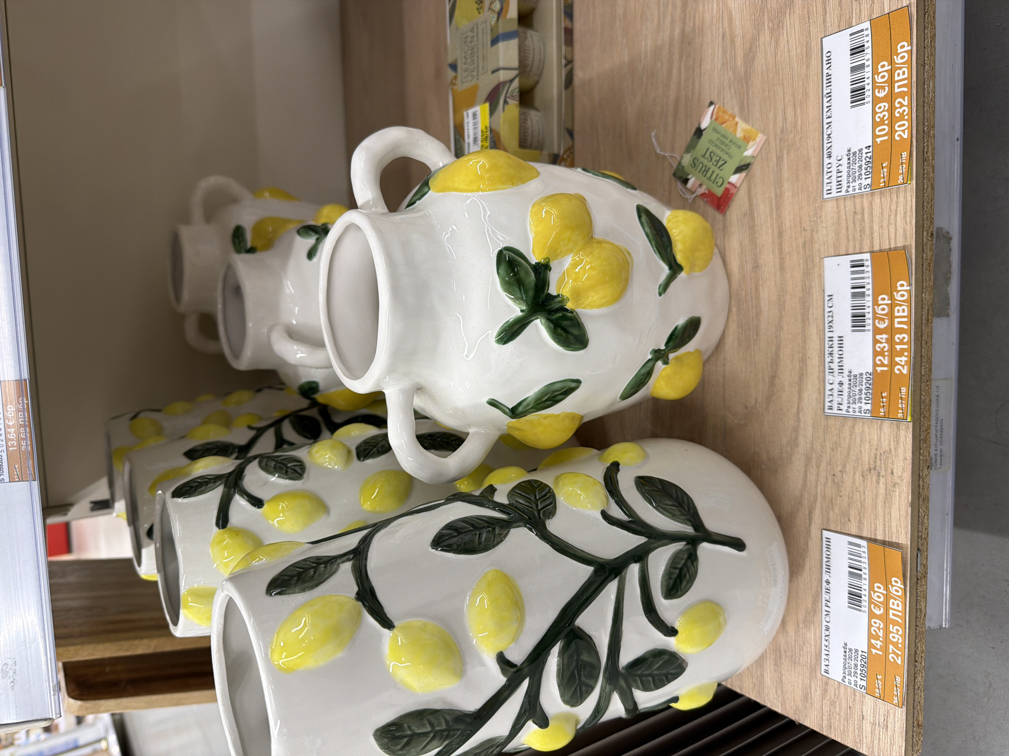 The image shows a retail display of ceramic vases decorated with lemon motifs. The vases are white with raised yellow lemons and green leaves. One large vase in the foreground is partially obscured by a smaller vase. There are price tags visible on the shelf beneath the vases. One tag reads "ВАЗА 15.5Х30 СМ РЕЛЕФ ЛИМОНИ" which translates to "VASE 15.5X30 CM RELIEF LEMONS". Another tag reads "ВАЗА С ДРЪЖКИ 19Х23 СМ ПЛАТО 40Х19СМ ЕМАЙЛИРАНО ЦИТРУС" which translates to "VASE WITH HANDLES 19X23 CM PLATFORM 40X19CM ENAMELED CITRUS". The price tags indicate prices in Euros and Bulgarian Leva. A small tag hanging from a vase reads "CITRUS ZEST FRAGRANCED CANDLE". The lighting suggests an indoor retail environment, possibly a home goods store. The background shows other merchandise and shelves. The specific location in the city of Plovdiv, Bulgaria, cannot be determined from the image alone.