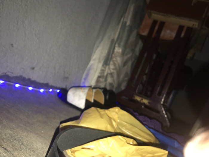 The image depicts an indoor scene in Kirana, Nigeria, under dim illumination. A string of active blue LED lights extends horizontally along the base of a light grey, textured wall. To the right of the lights, two partially rolled or folded white and black objects, possibly footwear, are situated. In the immediate foreground, a crumpled yellow plastic bag rests beside a dark object with a visible wide strap. In the background, a dark reddish-brown wooden or metal furniture frame, resembling a sewing machine stand, is partially visible against a patterned fabric backdrop. No individuals are present in the scene.