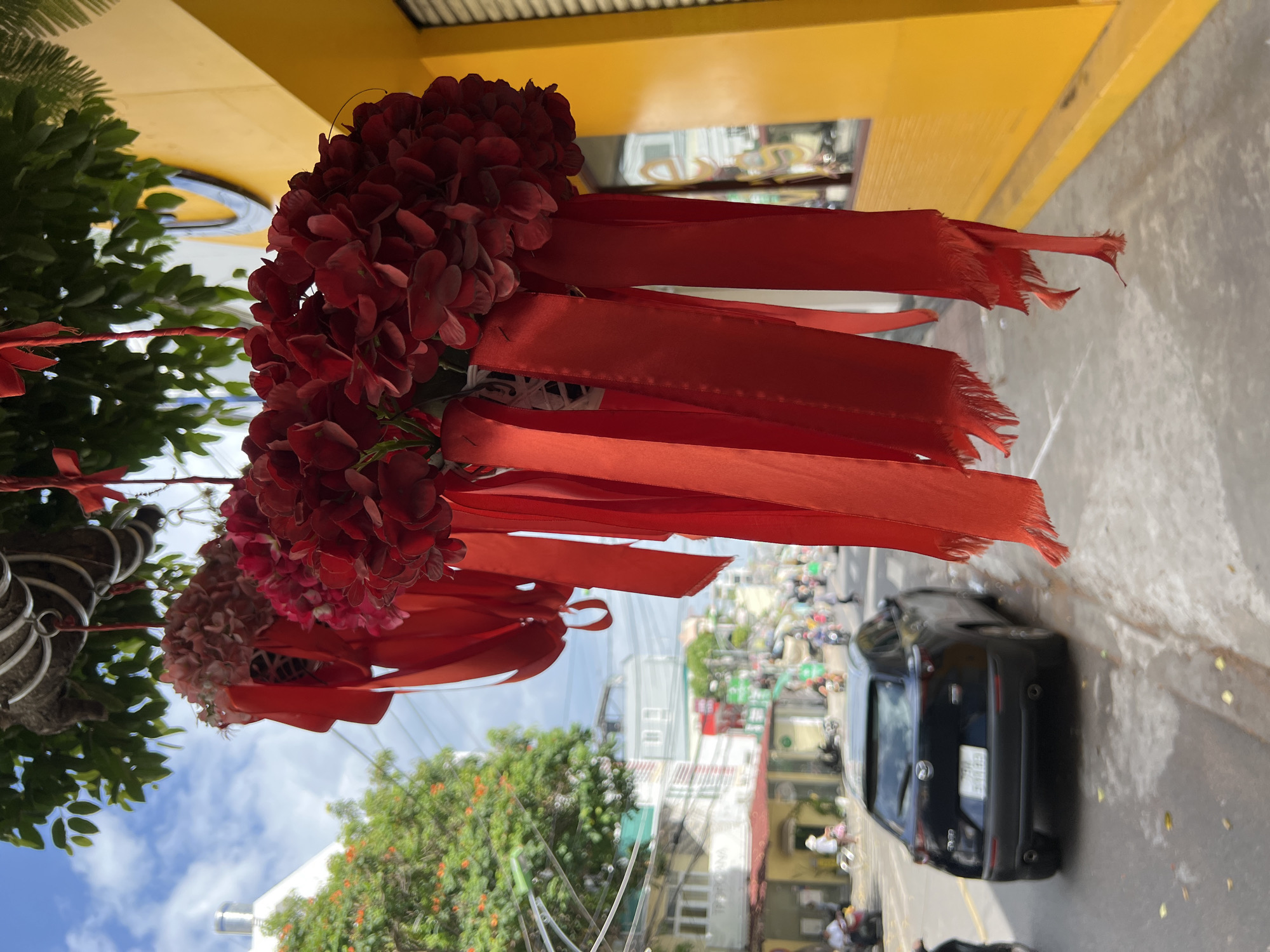 The image captures a street scene in Nha Trang, Vietnam, focused on decorative red floral arrangements with hanging red ribbons. These decorations are positioned in the foreground, obscuring a portion of the background. A black Mazda CX-5 is visible on the right side of the street, in the middle ground, with its rear facing the camera. The car is stopped at a crosswalk. A bright yellow building with a dark logo is on the right edge of the frame, with a window reflecting the street scene. The street itself appears wet, with reflections of the surrounding buildings and sky. In the background, there is a typical streetscape of buildings of varying heights, trees, and people on motorcycles, indicative of an urban environment in Vietnam. The sky is partly cloudy. The overall impression is one of a vibrant, bustling city street with decorative elements adding a touch of festivity.