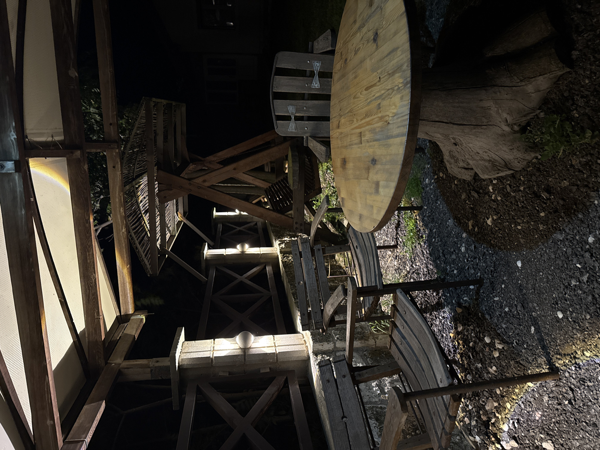 The image depicts a rustic outdoor seating area at night, situated in the coastal town of Tyulenovo, Bulgaria. A wooden table with a rough-hewn tree trunk base is centrally positioned, surrounded by several wooden chairs and benches. The seating is arranged on a gravel and dirt ground beneath a wooden and fabric canopy, which is illuminated by artificial lighting. In the background, a wooden swing set is visible, also appearing to be made of rustic materials. A wooden fence with crisscross patterns runs along the side of the area, with wall-mounted lights attached to stone pillars. The overall scene suggests an outdoor dining or relaxation space, likely part of a guesthouse or rural accommodation, given the natural and unrefined aesthetic. The lighting creates shadows, enhancing the nighttime ambiance. There are no people visible in the image.