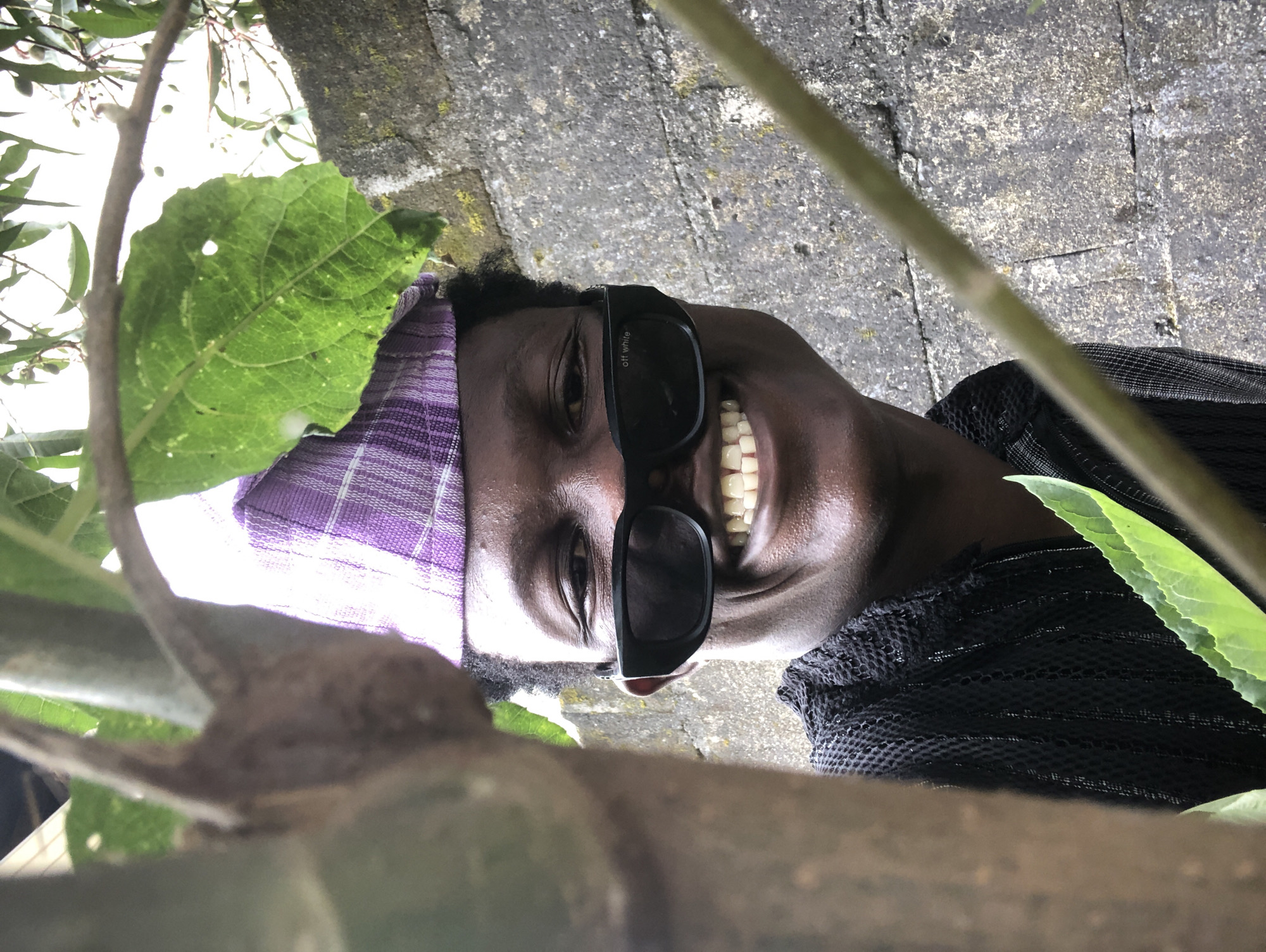 A young Black male is pictured outdoors, partially obscured by tree branches and leaves. He is wearing a purple and white patterned cap, black sunglasses with "off white" written on the left lens, and a black mesh shirt. He is smiling broadly, showing his teeth, and appears to be looking at the camera. The background consists of a textured, grey concrete wall and some greenery. The scene suggests a casual, possibly urban environment, consistent with locations in Ado, Nigeria.