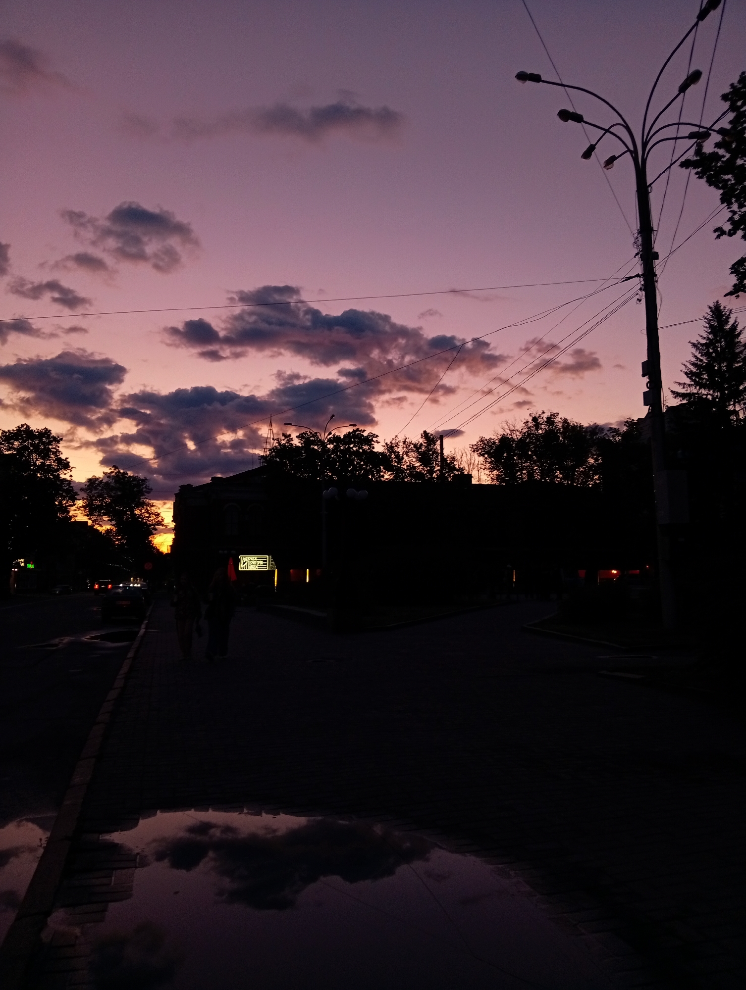The image depicts a twilight street scene in Poltava, Ukraine, marked by a vivid sky and silhouetted urban elements. The upper portion of the frame is filled with a stunning gradient of colors, transitioning from bright orange and yellow near the horizon to rich pink and deep purple overhead, interspersed with dark, scattered clouds.
In the mid-ground and foreground, the environment is largely rendered in silhouette. A prominent, multi-branched streetlight with several lamps stands on the right, its dark form stark against the colorful sky, accompanied by numerous overhead utility wires. Dark masses of trees, including what appears to be a coniferous tree, are visible along the skyline and in the background.
Below the horizon line, buildings and additional foliage are presented as dark silhouettes. On the left side of the street, two figures are faintly visible walking along the sidewalk, their forms obscured by shadow. Further down the street, subtle lights suggest the presence of vehicles. On a building in the mid-ground left, a lit sign is discernible, displaying the text "КОПІЙКА," a known supermarket chain in Ukraine.
A large puddle on the sidewalk in the bottom left corner reflects the dramatic sky and clouds above, hinting at recent rain. The overall lighting creates a serene yet moody atmosphere, emphasizing the contrast between the vibrant, glowing sky and the darkening urban landscape as day transitions into night.