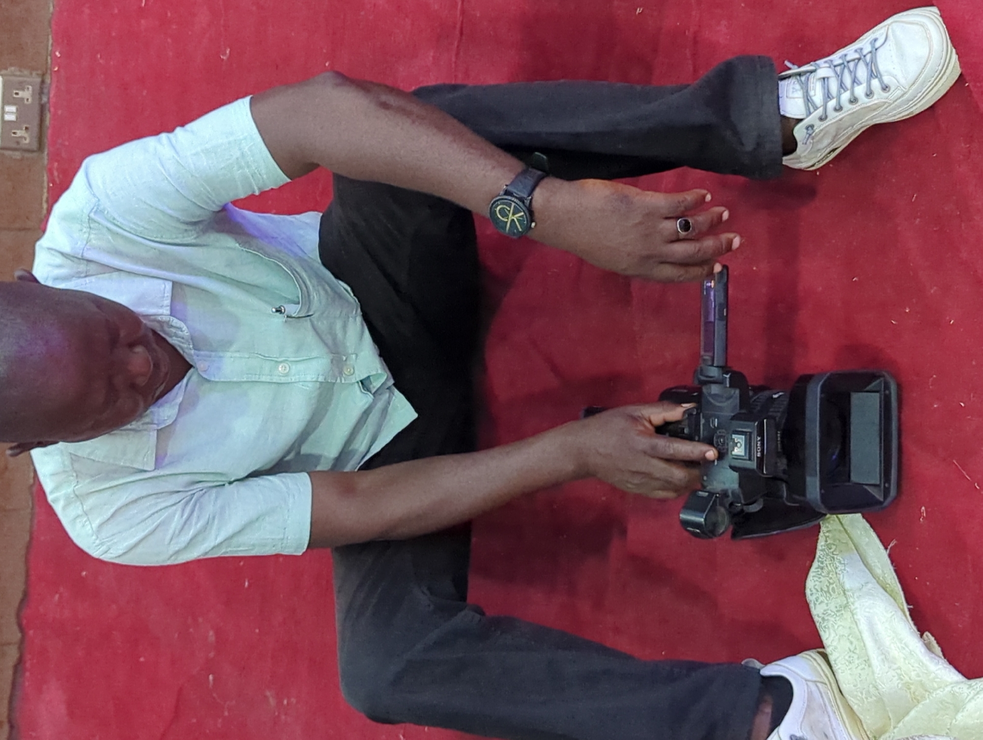 A person is sitting cross-legged on a red surface. The person is wearing black pants, a light-colored button-down shirt, a watch with a "CK" logo, and white sneakers with light blue laces. They are holding a black Sony video camera. Part of a white, patterned cloth is near the lower right corner. An electrical outlet is visible in the upper left corner. The location and city are not discernible from the image.