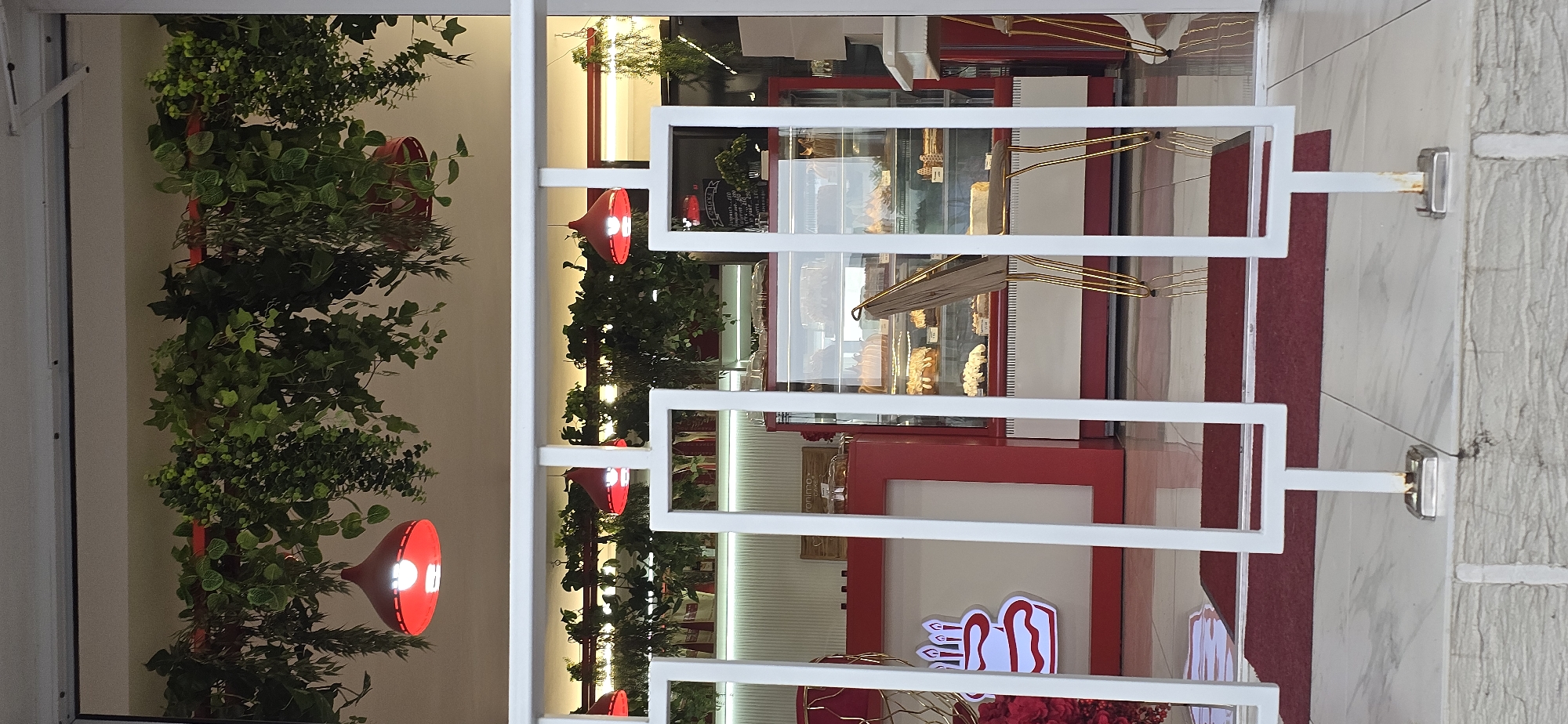 This is an image of a pastry shop viewed through a white modern gate. The gate has horizontal bars with a geometric design. On the left side of the image, there is an abundance of green foliage, possibly artificial plants, cascading down. Red lamps are interspersed among the plants, one of which is clearly visible and lit.
Through the gate, the interior of the shop is visible. It appears to be a well-lit space with red accents. There are display cases filled with baked goods, including cakes and pastries. The shelves and counters are predominantly red. The overall impression is of a clean and inviting establishment.
In the background, through the glass of the display cases, there are some reflections and hints of more items. There is some text visible on a black board behind the display cases, but it is too blurry to read. Further into the shop, there are more lights and decorative elements.
On the right side of the image, outside the gate, a textured light-colored wall is visible, with a red carpet running along the floor. The gate is attached to this wall with metal fixtures.
The lighting suggests it could be daytime, given the brightness of the interior and the illumination from the red lamps. The scene evokes a sense of a pleasant day for enjoying pastries.