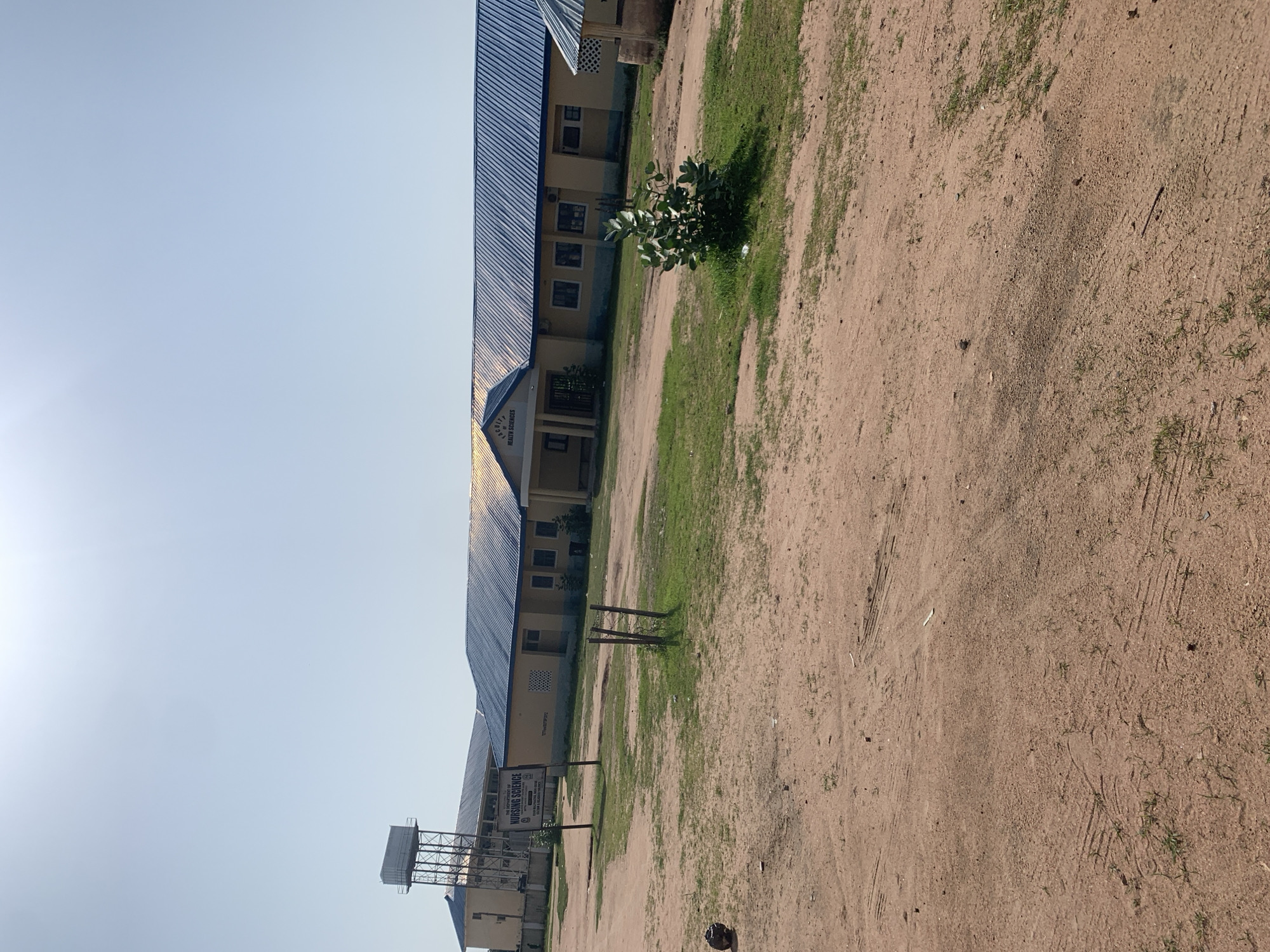 This is a wide-angle, eye-level, outdoor photograph taken on a clear, sunny day in Jalingo, Nigeria. The scene depicts a collection of buildings with blue, corrugated metal roofs, likely part of an educational or medical institution. In the background, a tall water tower with a rectangular tank is visible. A sign in front of one of the buildings reads "THE DEPARTMENT OF NURSING SCIENCE". Another building in the middle ground has a sign that says "FACULTY HEALTH SCIENCES". The foreground is dominated by a dirt and gravel area with patches of dry grass. Tire tracks are visible in the dirt. There are no people clearly visible in the image. The overall environment appears to be somewhat arid and rural, consistent with a location in Jalingo, Nigeria.