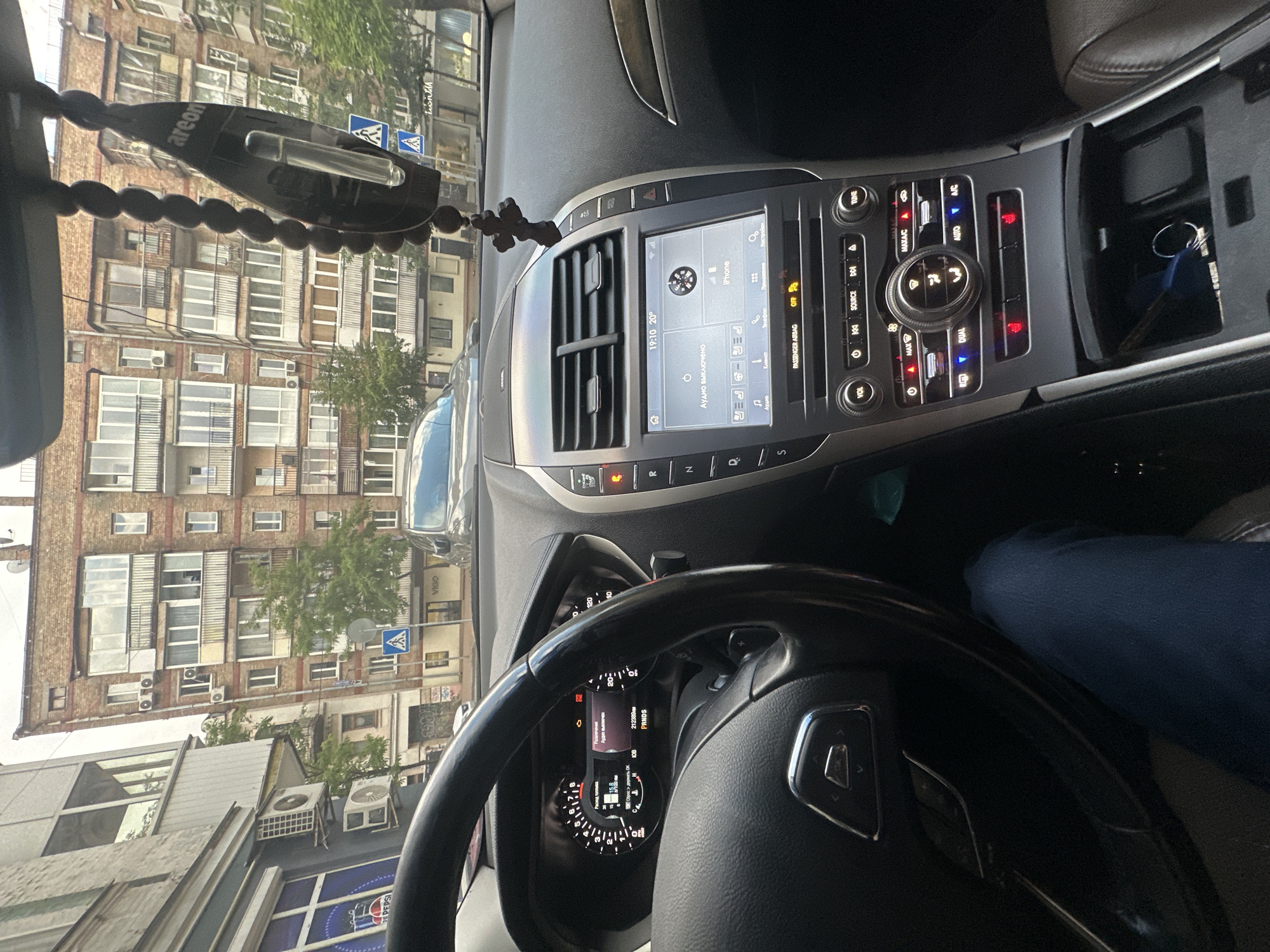 The image is a first-person perspective from inside a car, looking towards the dashboard and windshield, with a street scene in Kyiv, Ukraine, visible through the windshield. The dashboard features a digital instrument cluster on the left displaying speed and other vehicle information, and a central infotainment touchscreen on the right. The touchscreen displays the time as 19:10, the temperature as 20 degrees Celsius, and shows that audio is currently muted ("АУДИО ВЫКЛЮЧЕНО"). An iPhone is connected and displayed on the screen. A black steering wheel with silver accents is visible in the foreground, and a person's leg in blue jeans is also visible, suggesting the photographer is the driver. A car air freshener, a string of wooden beads with a black pendant, is hanging from the rearview mirror. Outside the car, a multi-story brick building with balconies and many windows is prominent. Trees and street signs are also visible on the street. The overall impression is of a vehicle parked or stopped on a city street.