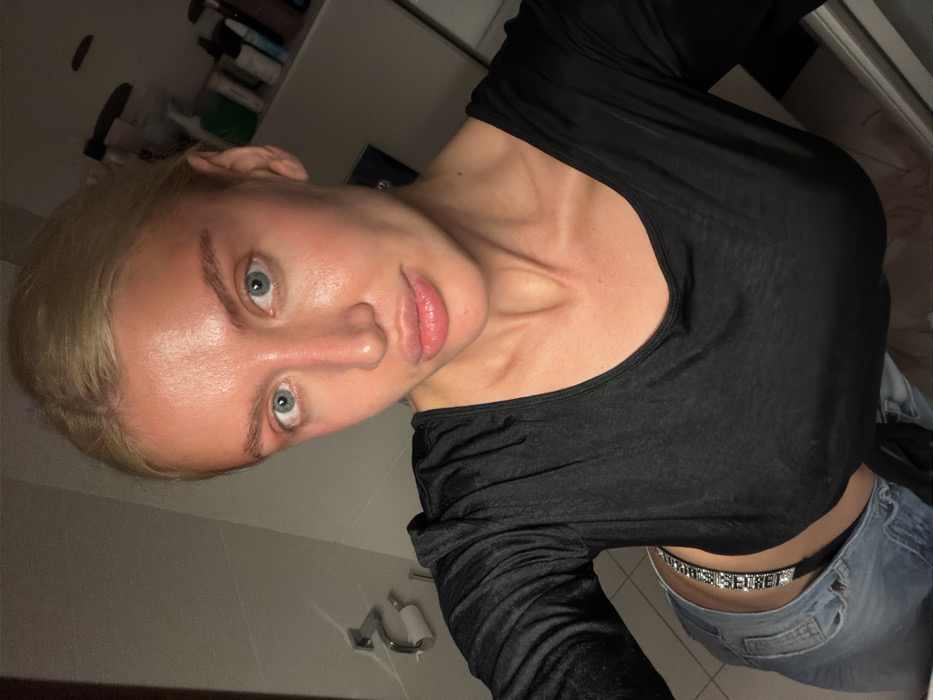 The image shows a young adult female taking a selfie in a bathroom located in Budva, Montenegro. She has blonde hair pulled back, blue eyes, and full lips. Her skin, particularly on her face, neck, and upper chest, exhibits significant erythema consistent with sunburn. She is wearing a black, long-sleeved, scoop-neck top and light blue denim jeans. A silver-toned, possibly rhinestone-studded belt or waist accessory, spelling "SISTER," is visible at the waistband of the jeans. The background on the left includes a chrome toilet paper holder with a white roll mounted on a light-colored wall. To the right, a shelf or counter displays various personal care bottles and containers. The scene is illuminated by artificial, slightly dim lighting.