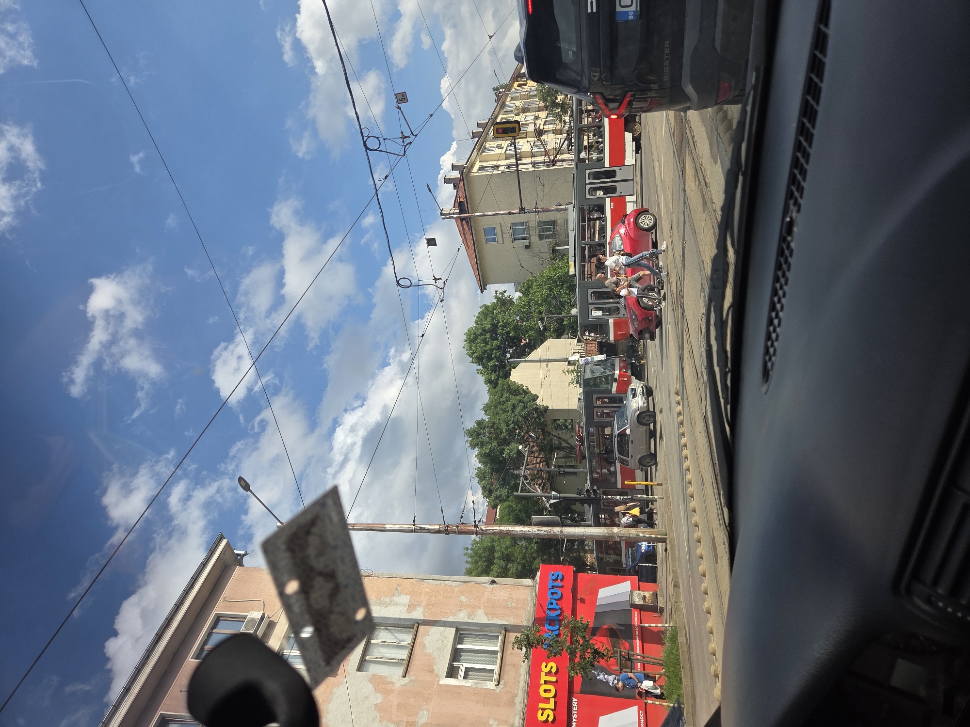 This image captures a vibrant urban street scene in Sofia, Bulgaria, viewed from inside a vehicle, with parts of the dashboard visible on the right. The scene is oriented vertically in the original, but describes a horizontal streetscape.
The primary subjects include several people, various vehicles, and urban infrastructure under a bright, partly cloudy sky. A red and white tram runs along tracks in the center-left. Next to the tram, a small red car is stopped, surrounded by a few individuals. Two people are mounted on a scooter or moped near the red car, seemingly interacting with the group. Further down the street, other vehicles and pedestrians are visible.
Buildings line both sides of the street. On the left, a multi-story building with a weathered facade and exposed wires is prominent, with a large red sign at its base. On the right, a yellow-beige building with numerous windows and balconies rises behind the tram lines. Overhead, a dense network of power lines for the tram system crisscrosses the blue sky, punctuated by streetlights and utility poles.
The activity suggests a normal daytime scene with ongoing traffic and pedestrian movement. The specific interaction around the red car and scooter might indicate a brief stop or a casual gathering.
Notable details include the sunny weather with scattered white clouds. A Bulgarian license plate ("BG" visible) on a black SUV partially seen in the top right confirms the location. The red sign prominently displays "SLOTS JACKPOTS" in large white letters, with other smaller, unreadable text below. A rectangular, blurry object, possibly a sticker or permit, is adhered to the inside of the camera vehicle's windshield on the left. The time of day is clearly daylight.