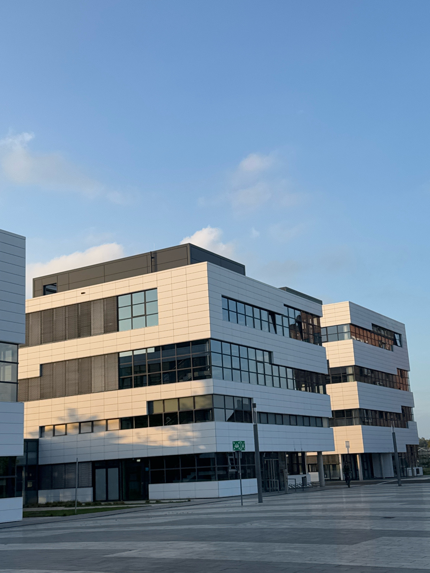 The image shows a modern office building complex in the city of Cleves, Germany. The building features a contemporary architectural design with white and light-colored facade panels, interspersed with large glass windows and dark-colored horizontal blinds. The structures are multi-story and appear to be connected or part of a larger complex. The sky above is a clear blue with a few wispy clouds, indicating daylight. In the foreground, a paved area, possibly a plaza or walkway, leads up to the buildings. A single person is visible on a bicycle in the middle ground, riding away from the viewer. The overall scene suggests a business or corporate environment, with the architecture reflecting a modern aesthetic.