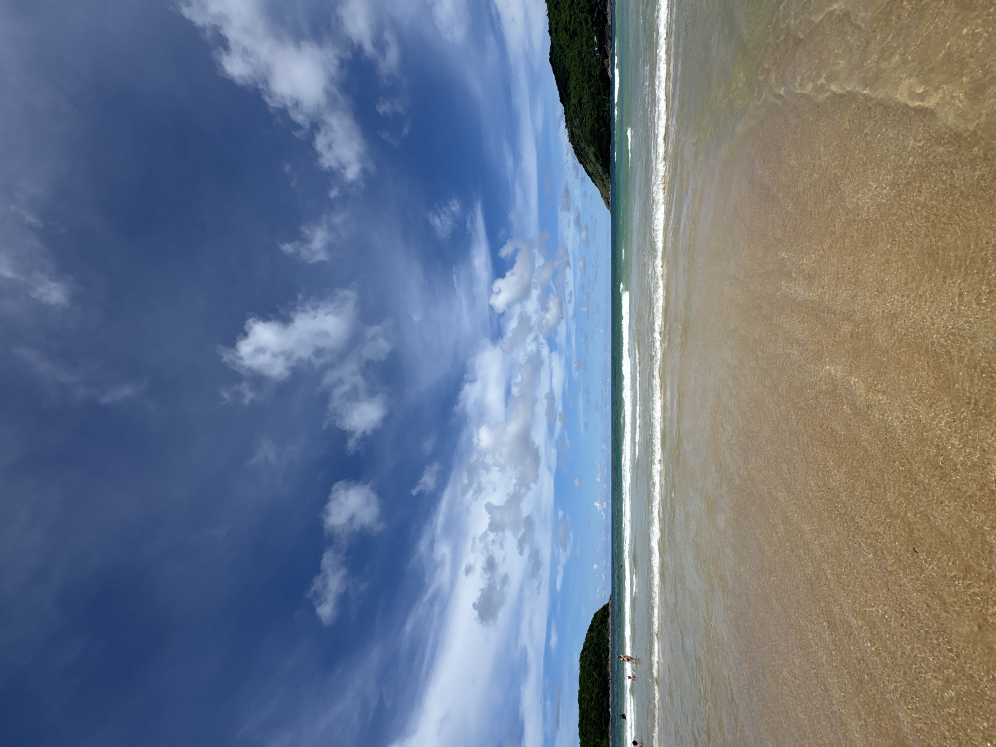 The image captures a picturesque beach scene in Rawai, Thailand, under a bright, partly cloudy sky. The foreground is dominated by the wet sand of the beach, with the shallow, clear water gently lapping at the shore, revealing the textured sandy bottom. In the mid-ground, small waves with white crests roll towards the beach. A few people are visible in the water, engaged in leisure activities. Two small, tree-covered islands flank the horizon on either side, with the larger island on the right being more prominent. The turquoise ocean stretches towards the horizon, meeting the deep blue sky dotted with white, wispy clouds.