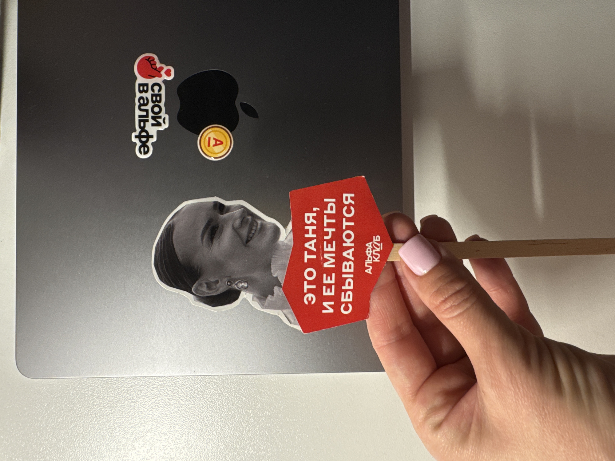 A hand holds a red sign that reads "ЭТО ТАНЯ, И ЕЕ МЕЧТЫ СБЫВАЮТСЯ," which translates to "THIS IS TANYA, AND HER DREAMS COME TRUE," next to a laptop. The sign includes the text "АЛЬФА КЛУБ" which translates to "ALPHA CLUB." The laptop has stickers of a heart-shaped hand with the text "СВОЙ ВАЛЬФЕ" which translates to "YOUR VALFE" and an image of the letter "A" in a yellow circle. A black and white sticker of a woman's face is also affixed to the laptop. The background is a white surface. There is no specific information to definitively identify the location as Sochi, Russia.