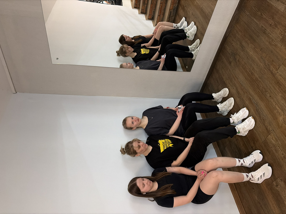 Three young adult females are depicted indoors, performing a wall sit exercise in a facility located in Chaikovsky, Russia. The subjects are positioned side-by-side against a plain white wall. From left to right:
1. A female with long, dark brown hair and a white earbud in her left ear, wearing a black short-sleeved top, black shorts, and white athletic shoes with patterned socks. Her head is angled downwards.
2. A female with blonde hair tied in a bun, wearing a black short-sleeved t-shirt featuring a yellow graphic with the text "PRIDE SPORT FEST," black athletic pants, and white athletic shoes. Her head is angled downwards.
3. A female with long, light blonde hair, wearing a dark grey short-sleeved t-shirt, black leggings, and light grey athletic shoes. She holds a white smartphone in her left hand and maintains a neutral facial expression, looking forward.
All three individuals are in a wall sit posture, with their backs pressed against the white wall and their knees bent at approximately 90 degrees, simulating a seated position without a chair. Their feet rest flat on a dark brown wooden floor. The right side of the frame features a large, rectangular mirror, reflecting portions of the subjects, a light grey wall corner, and a brown wooden staircase with exposed brick-like risers in the background.