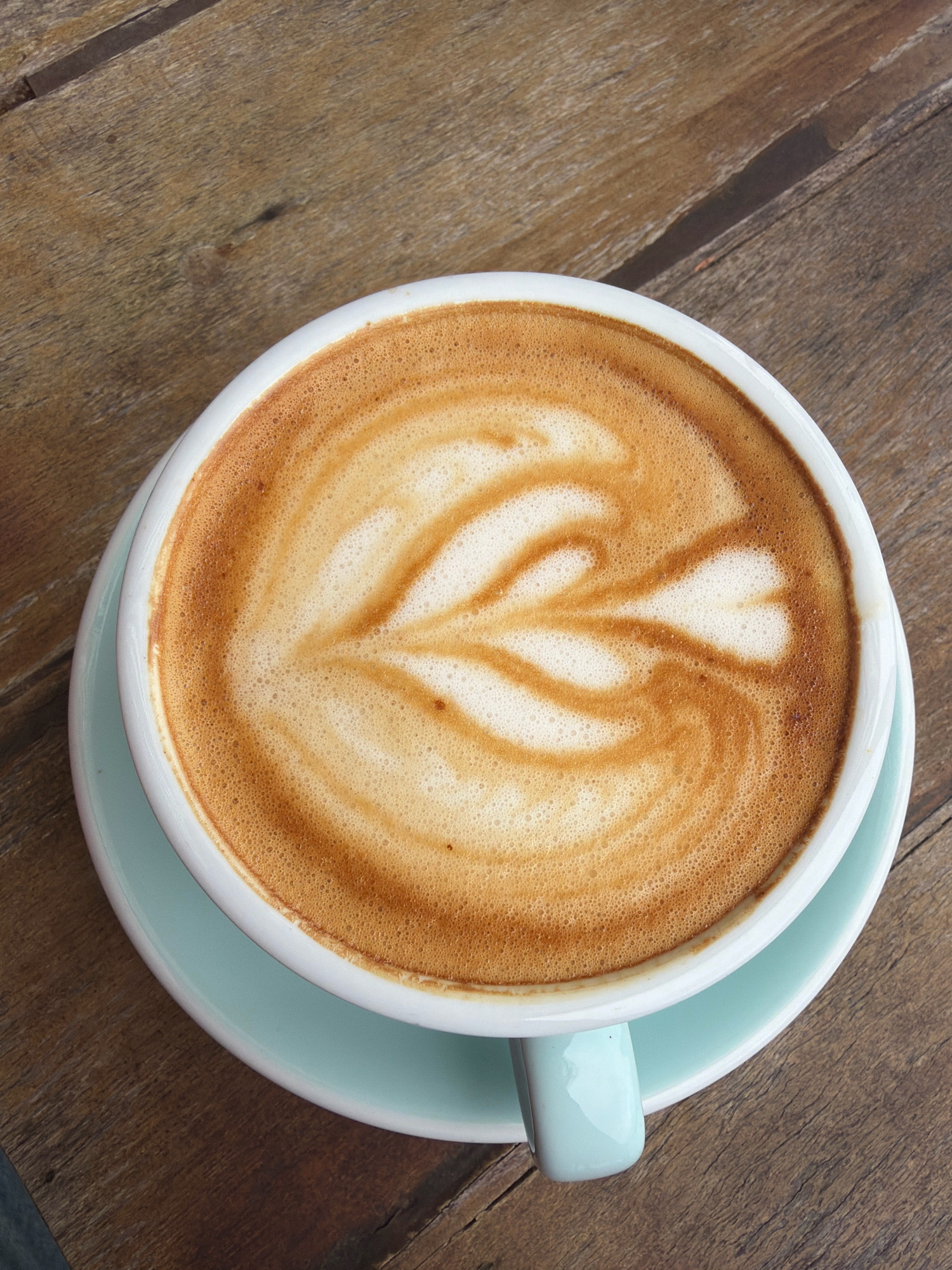 A close-up, overhead shot captures a ceramic mug of latte with intricate foam art resting on a light blue saucer, all placed on a rustic wooden table. The latte features a delicate pattern of swirling milk foam against the rich brown coffee. The mug and saucer are a pale blue, contrasting with the warm tones of the coffee and the dark, textured wood grain of the table. The setting does not provide any specific clues to its location within Da Lat, Vietnam.
