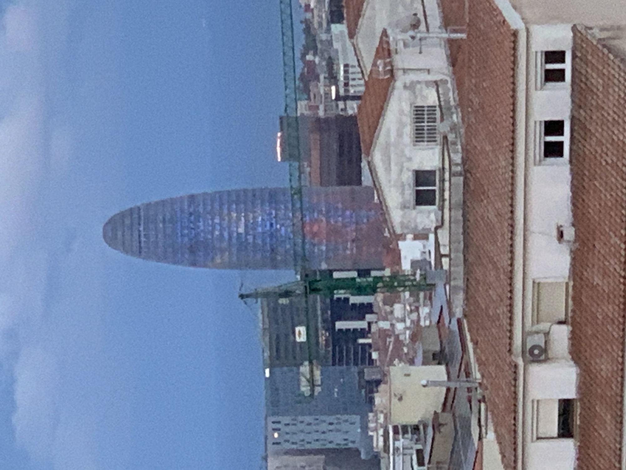The image depicts a cityscape with the Agbar Tower prominently featured. The tower, a distinctive bullet-shaped skyscraper, dominates the skyline with its colorful patterned facade. A construction crane is visible near the tower, suggesting ongoing building activity in the area.
Adjacent to the tower are older, low-rise buildings with terracotta roofs and white facades. The presence of residential features such as windows and air conditioning units on these buildings indicates a mix of old and new architecture in the urban environment.
The sky in the background is a gradient of blue, suggesting either dusk or dawn. The image captures a typical view of the city, showcasing its modern landmarks alongside its traditional architecture.