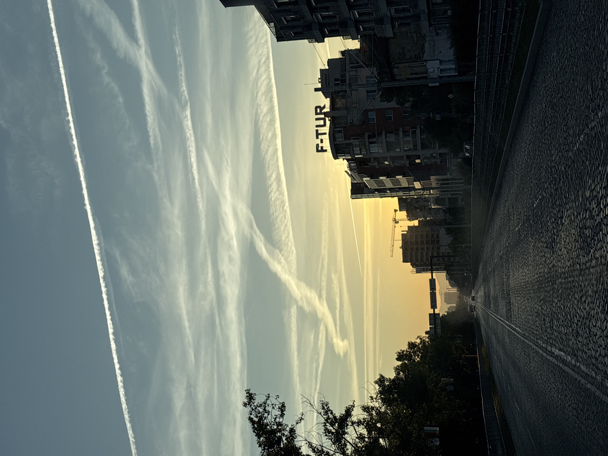The image shows a view of a city street at sunrise or sunset in Sofia, Bulgaria. The foreground is a cobblestone road with lane markings, leading into the distance where several cars are visible. On either side of the road are trees and buildings. The buildings vary in height and architectural style, with one prominent building featuring the illuminated sign "F-TUR." Construction cranes are visible in the background, suggesting ongoing development. The sky is a gradient of blue and yellow, with numerous contrails crisscrossing. The overall impression is an urban landscape captured during a transitional time of day, with signs of both established infrastructure and ongoing construction.
