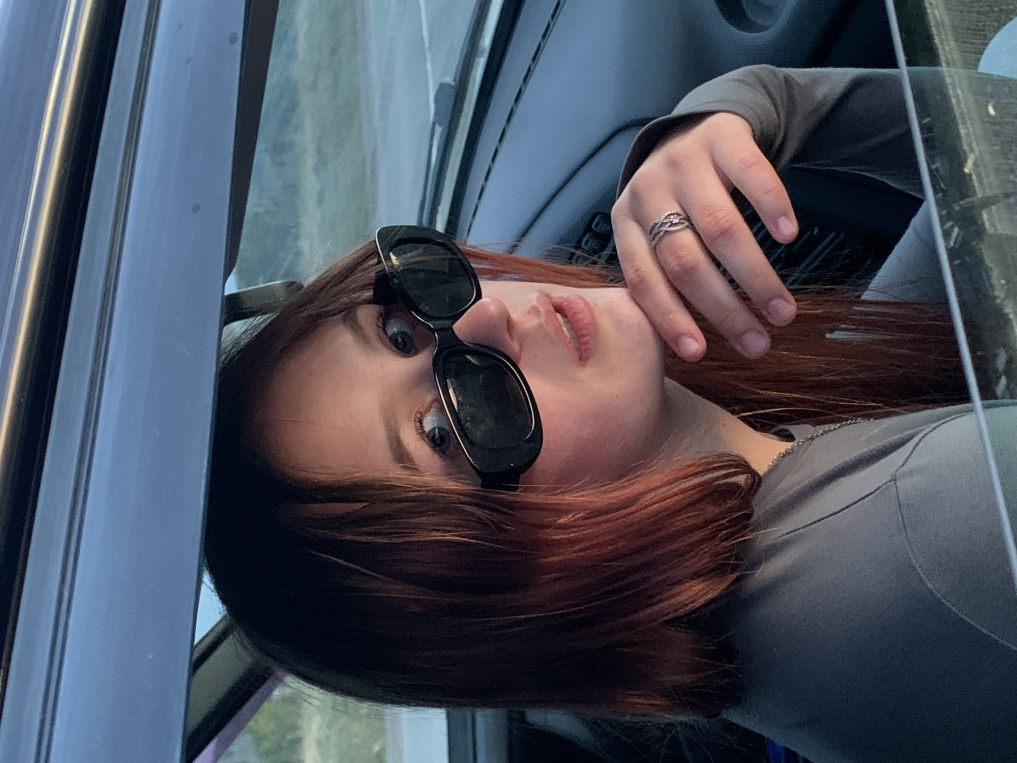 A close-up, rotated portrait shows a young person with reddish-brown, shoulder-length hair, situated inside a vehicle. The individual, appearing to be female, is wearing dark, rectangular sunglasses and a grey long-sleeved shirt. Her left hand, adorned with an intricately designed, possibly silver-colored ring on her ring finger, is gently raised, with her fingers touching her chin. She looks directly towards the camera with a neutral expression.
The setting appears to be a car interior, with parts of the car door frame, window, and dashboard visible around her. Through the car window on the right side of the image, a blurry outdoor scene suggests a natural or distant landscape. The lighting is soft and even, indicating daytime, possibly late afternoon or early evening. There are no explicit weather conditions visible. No text is visible in the image. The location context provided for this image is Фоки, Russia.