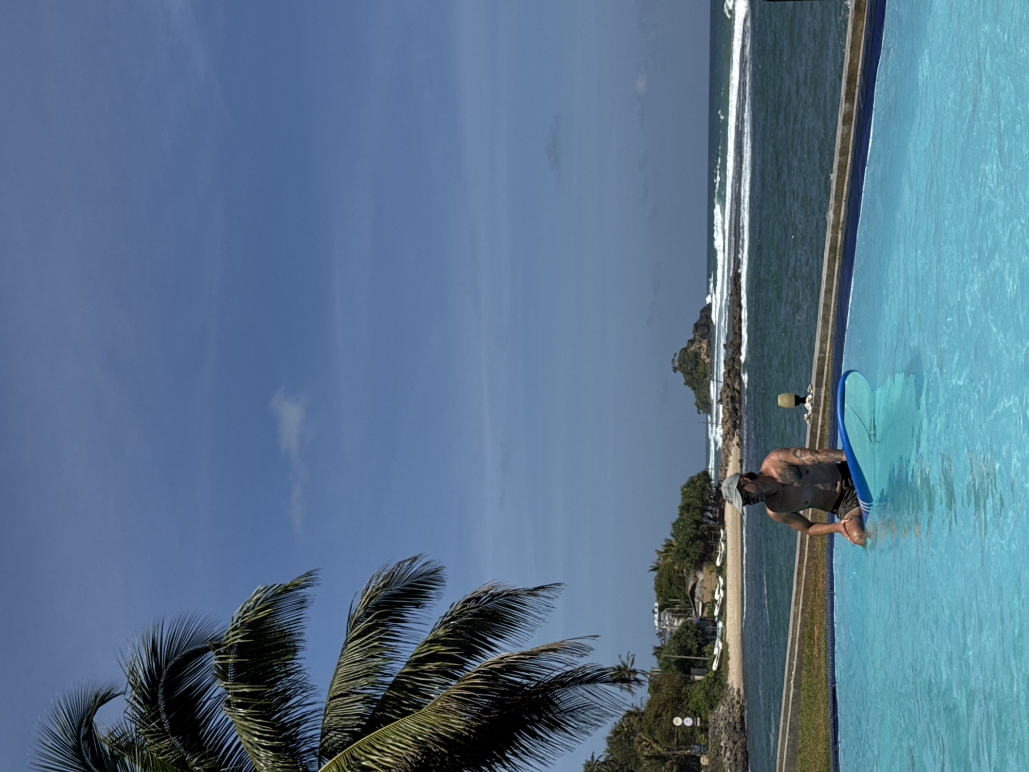 A man wearing a grey cap and dark shorts sits on a surfboard in a bright blue swimming pool. He is shirtless and has tattoos on his arms and chest. The pool is in the foreground, with the ocean visible in the background. The ocean is dark blue with white waves and a small, rocky island with trees in the distance. To the left of the pool, a palm tree with green fronds extends into the upper left corner of the frame. Behind the ocean, there is a sandy beach lined with trees and a few buildings. The sky is clear and blue with a few small white clouds. The scene appears to be a resort or hotel in Goviyapana, Sri Lanka, given the tropical vegetation and coastal setting.