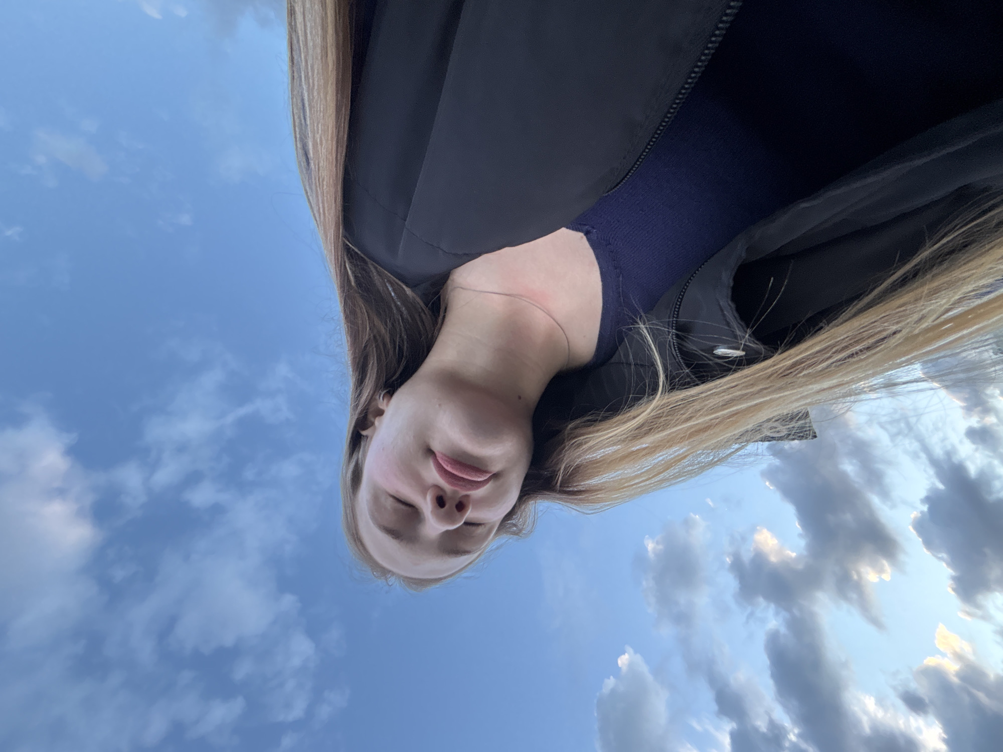 A low-angle, medium shot captures a young person with their eyes closed, tilted slightly upwards against a blue sky with scattered clouds. Their face is turned to the side, with blonde hair falling across their shoulder. They are wearing a navy blue shirt and a black jacket. A thin silver necklace is visible around their neck. The location is implied to be Solnechnoye, Russia, suggested by a contextual element not present in the visual data itself. The overall mood is serene and contemplative, with the focus on the person's peaceful expression and the expansive sky.