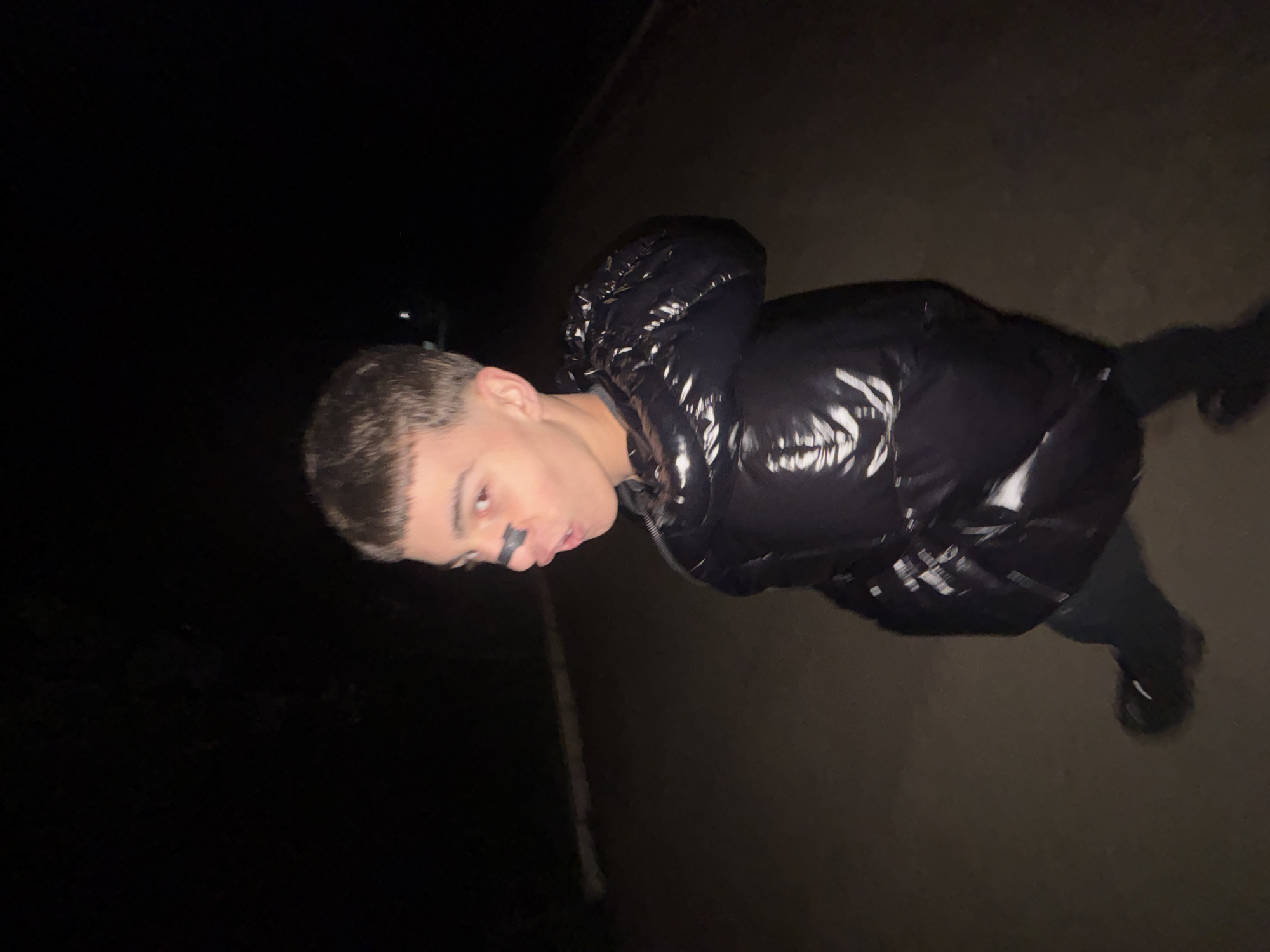 A person wearing a shiny black puffer jacket with the hood up stands in the dark. The person's face is visible and they have a dark mark on their upper lip. The background shows a dark road or sidewalk. The location is unspecified.