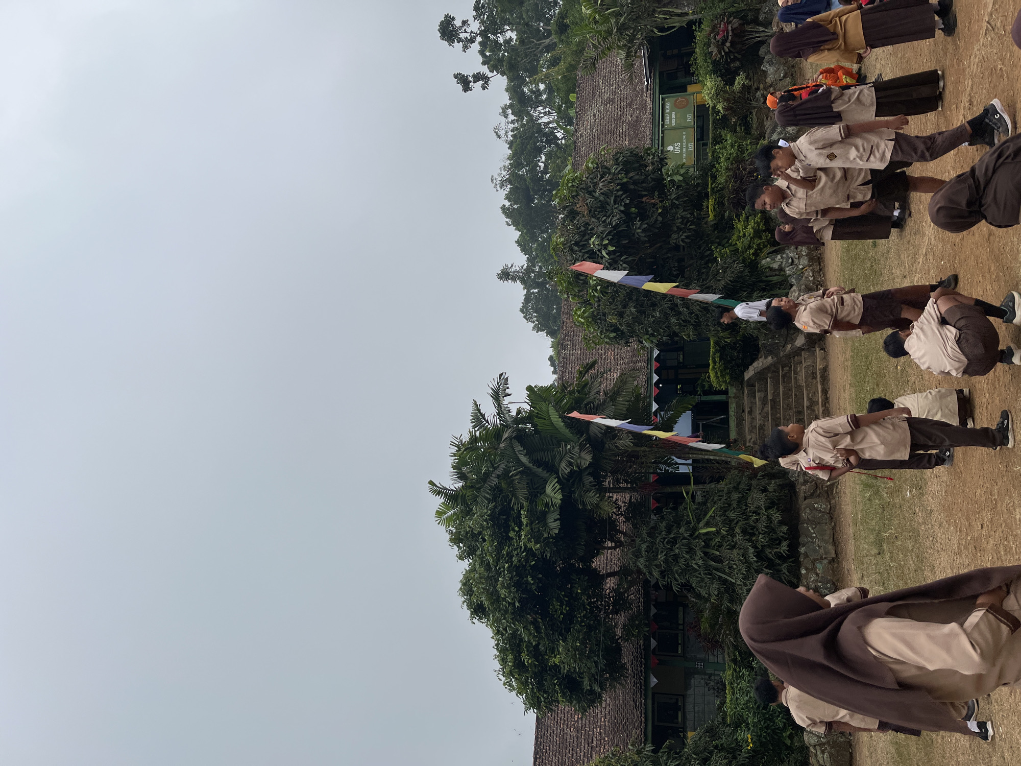 A group of students in uniform are gathered in a schoolyard in Gembong, Indonesia. The students, dressed in light brown shirts and darker brown pants or skirts, are standing and interacting in small groups. One student, wearing a brown hijab, is in the foreground looking to the left. Another student, also in a brown hijab, is in the far foreground with their back to the camera. Behind the students is a single-story school building with a tiled roof, flanked by lush green trees. Several flags, including a red and white flag, are visible near the building. The sky is overcast and the ground is a mixture of dirt and grass. A sign on the building reads "UKS" and lists an address in Pati, which is the regency Gembong is located in. The overall scene suggests a school event or a break period.