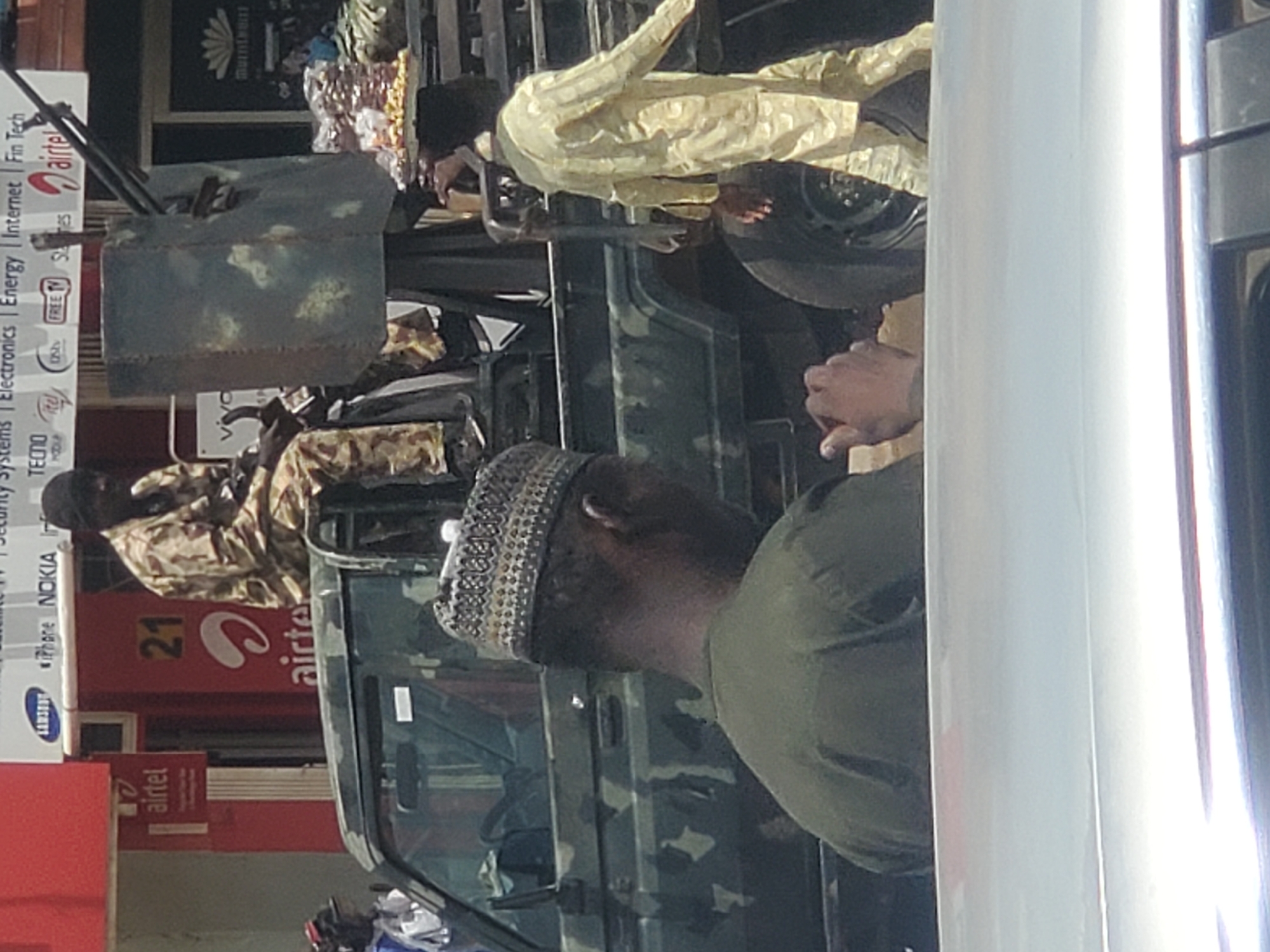 The image captures a street scene in Jos, Nigeria, showing a camouflaged military vehicle with several individuals. One man, wearing a military uniform, is seated in the back of the vehicle, while another man, wearing a traditional white and yellow robe, stands beside it. A third man, wearing a cap and green shirt, stands in the foreground. The backdrop includes storefronts with visible advertisements for brands like Airtel, Samsung, Nokia, Tecno, and Itel.