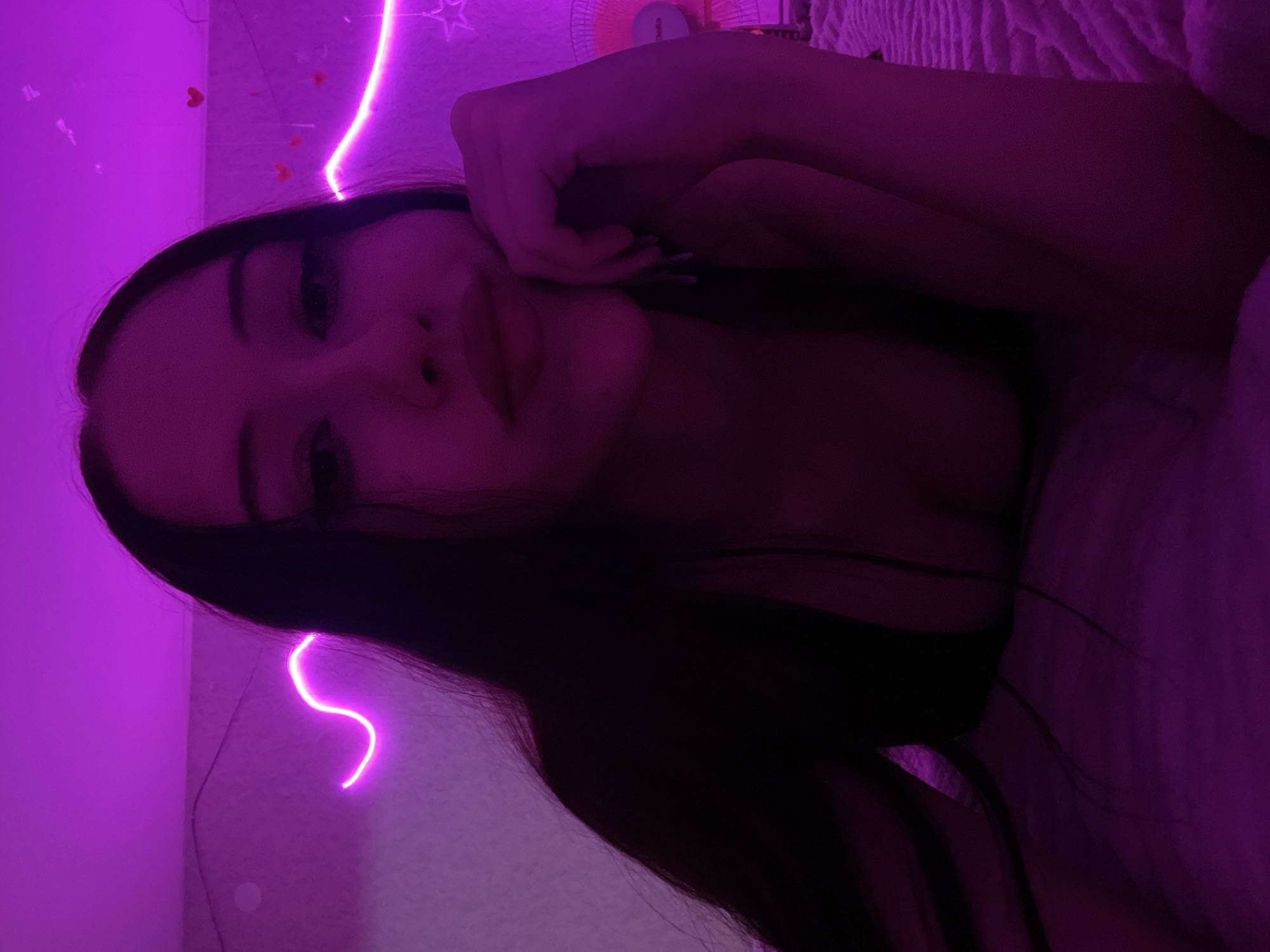 The image depicts a young woman with long, dark hair lying on a textured surface, possibly a bed, under purple ambient lighting. She is looking towards the camera with her chin resting on her hand. The background features decorative neon lights, including a wavy line and some heart-shaped elements. There are also some faint outlines of objects in the background, but they are not clearly discernible due to the lighting and focus. The woman is wearing a dark top that reveals a portion of her cleavage. The overall impression is that of a selfie taken in a dimly lit room with decorative lighting. The location of Votkinsk, Russia, is not discernible from the image itself.