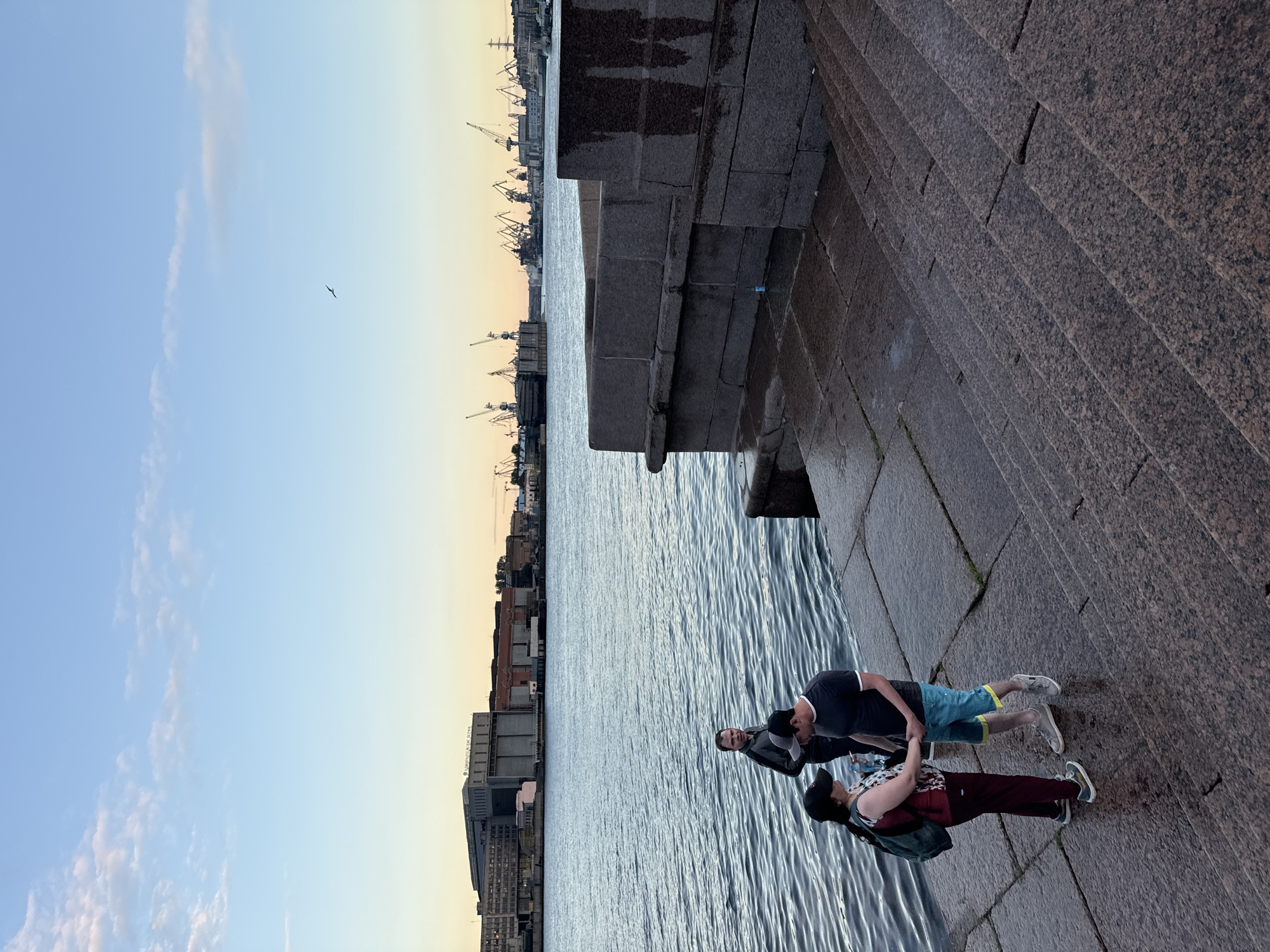 The image depicts a waterfront scene in Saint Petersburg, Russia, likely during the late afternoon or early evening given the soft, warm light and clear sky. Three individuals are present on a granite embankment by the water. In the foreground, a couple stands holding hands, facing each other. The woman wears a dark baseball cap, a patterned top, and dark leggings with a backpack. The man, also wearing a baseball cap, is dressed in a dark t-shirt and colorful shorts. Behind them, a third individual stands looking towards the water.
The embankment itself is constructed from large, dark granite blocks, with steps leading down to the water's edge, which appears wet. The water is calm and reflects the sky and distant industrial elements. Across the water, the Saint Petersburg skyline is visible, characterized by a large, multi-story industrial building with many windows and signage. Further in the distance, a series of large cranes and industrial structures are silhouetted against the horizon, suggesting a port or shipyard. The sky is a pale blue, transitioning to a warmer hue near the horizon, with a few wispy clouds. A single bird is captured in flight against the sky. The overall impression is one of a peaceful, urban waterfront setting.