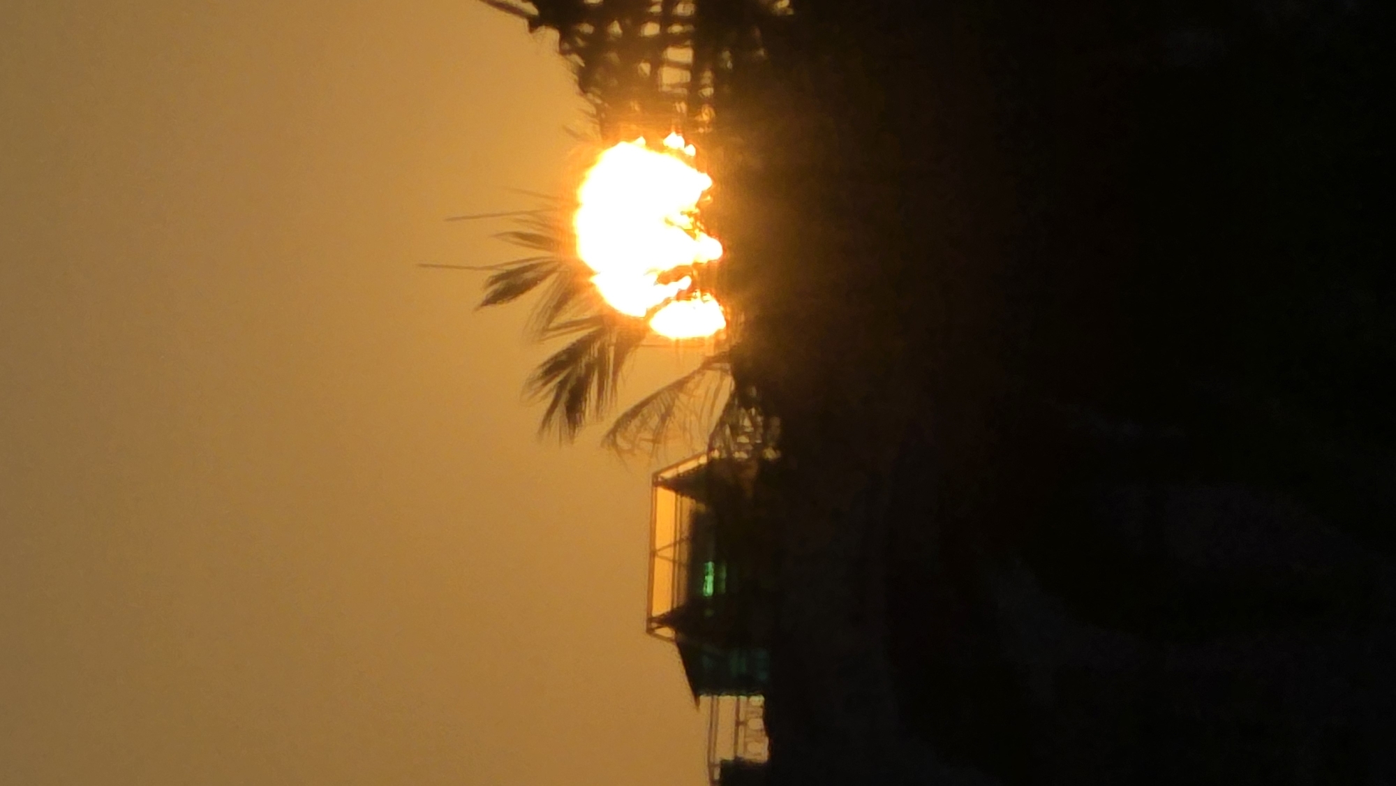 The image captures a sunset in Nha Trang, Vietnam. The sun, a bright orange-yellow orb, is partially obscured by palm tree fronds. A building with a bright yellow balcony and a green window is silhouetted against the sun. The sky has an orange-yellow hue, and the rest of the image is shrouded in darkness, particularly to the right side, suggesting the time of day.