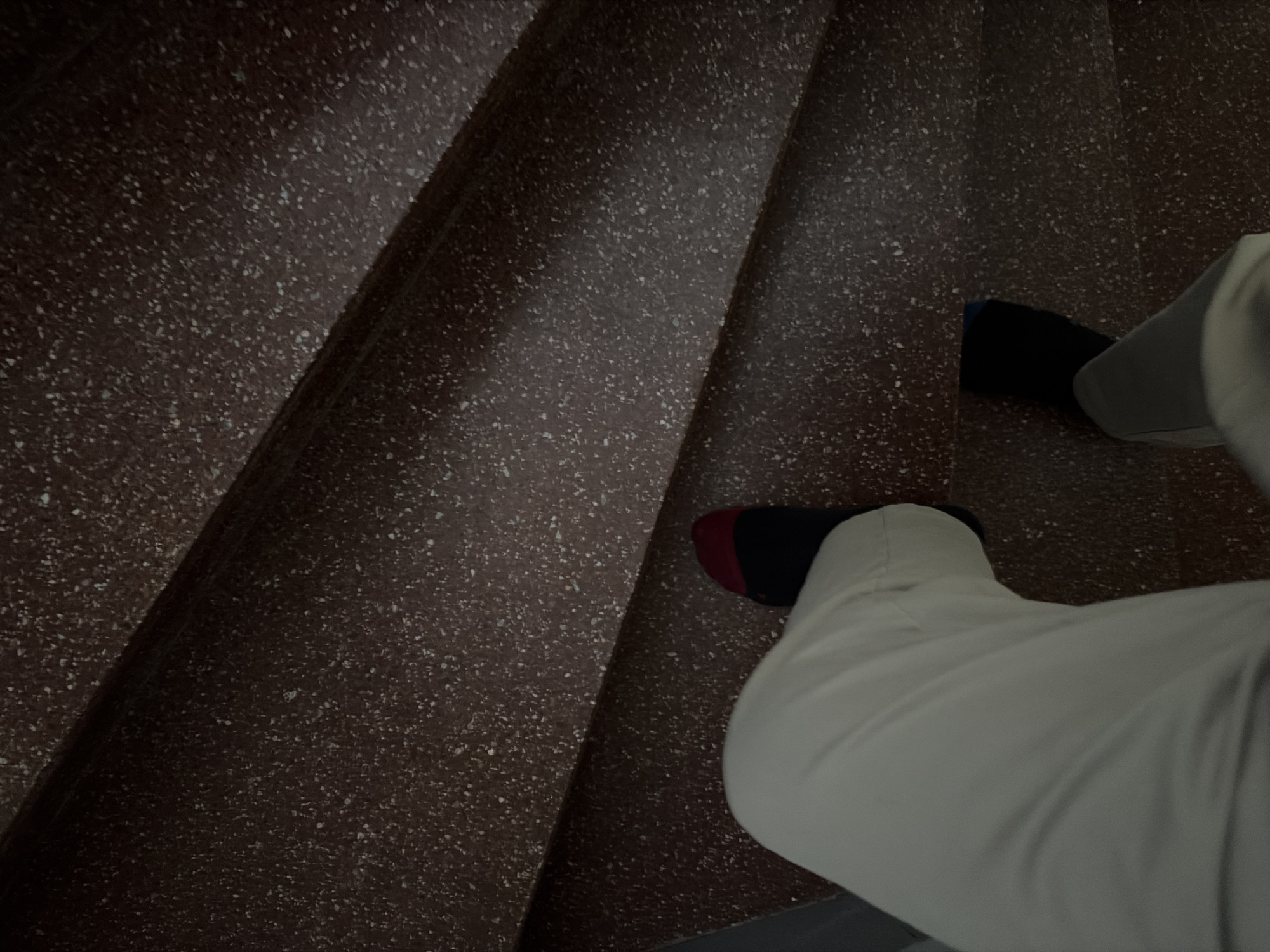 The image depicts a downward view of a person walking down a flight of stairs. The stairs are covered in a brown speckled material. The person is wearing light gray pants and socks of different colors: one sock is predominantly red with a black toe and heel, while the other is predominantly black with a blue toe and heel. The scene is located in Pétange, Luxembourg.