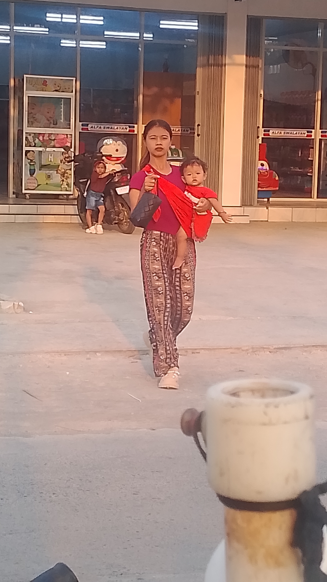 A young woman is walking with a baby slung on her shoulder in front of a row of shops in Karanglangu, Indonesia. The woman is wearing a magenta top and patterned trousers. The baby is wearing a red top. In the background, there is a motorcycle with a child sitting on it. The shops have signs that say "ALFA SWALAYAN". The ground is paved.