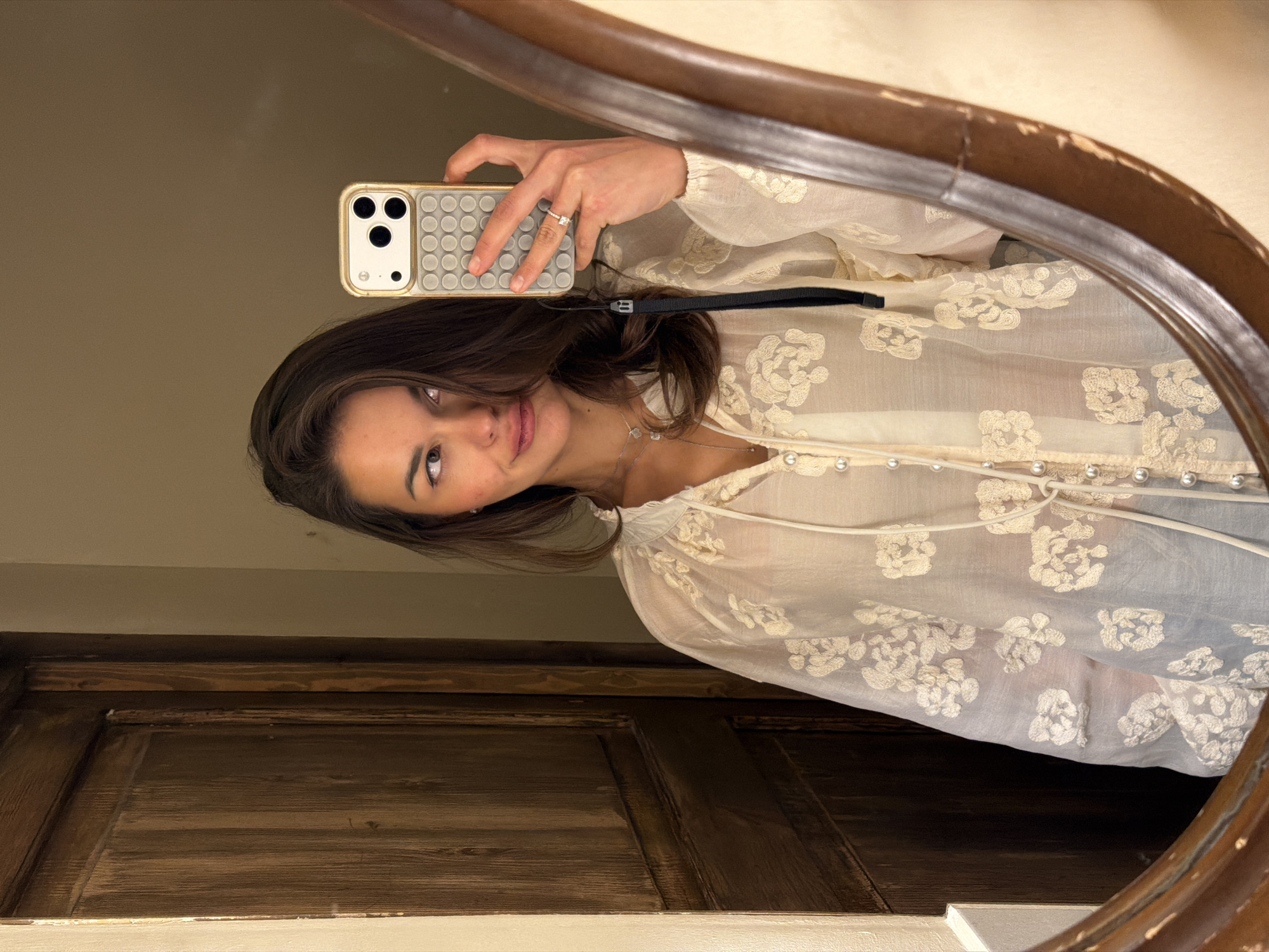The image depicts a female subject taking a mirror selfie. She has dark brown hair parted on the right, light-colored eyes, and a subtle smile, with her gaze directed toward the phone screen. She wears a sheer, cream-colored blouse featuring floral embroidery, a v-neck with a tie, and a front closure with small pearl-like buttons. Two silver-toned necklaces are visible, along with a stud earring in her right ear and a ring with a clear stone on her left ring finger. She holds a gold-colored smartphone with a light grey, circular-patterned (pop-it style) case and a black wrist strap in her right hand. The mirror has a dark brown, possibly wooden, frame showing signs of wear along its edges. Reflected in the mirror is a textured dark wooden door on the left and a plain light beige wall on the right side of the subject. No specific visual cues are present to identify a location within Tbilisi, Georgia.