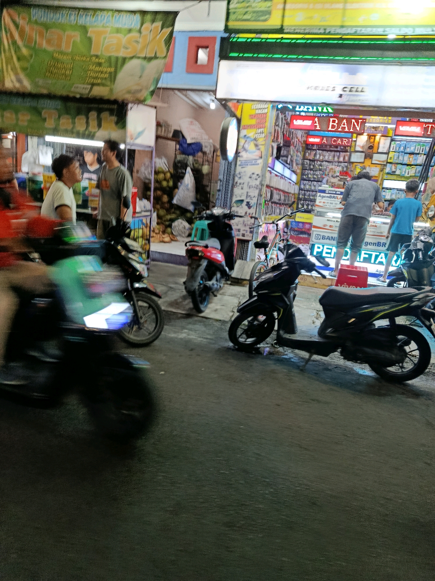 This is a street-level, slightly blurry, nighttime photo taken in Bekasi, Indonesia. In the foreground, two scooters are parked, one black and one mostly out of frame but appears to be green and white. A third scooter, black with yellow accents, is in the mid-ground.
In the mid-ground, behind the scooters, is a street-side stall or shop. A sign above the stall reads "KELAPA MUDA" which translates to "young coconut". Another sign, partially obscured by the scooters, appears to advertise "Tasik".
Further back, a brightly lit electronics store or mobile phone shop is visible. Signs in red and blue neon lights read "ALES CELL" and "A BAN". Several people are seen inside and in front of the shop, including a person wearing a grey shirt and shorts, and another in a blue t-shirt. Products, likely mobile phones and accessories, are displayed on shelves inside the shop. The overall scene depicts a busy commercial area at night.