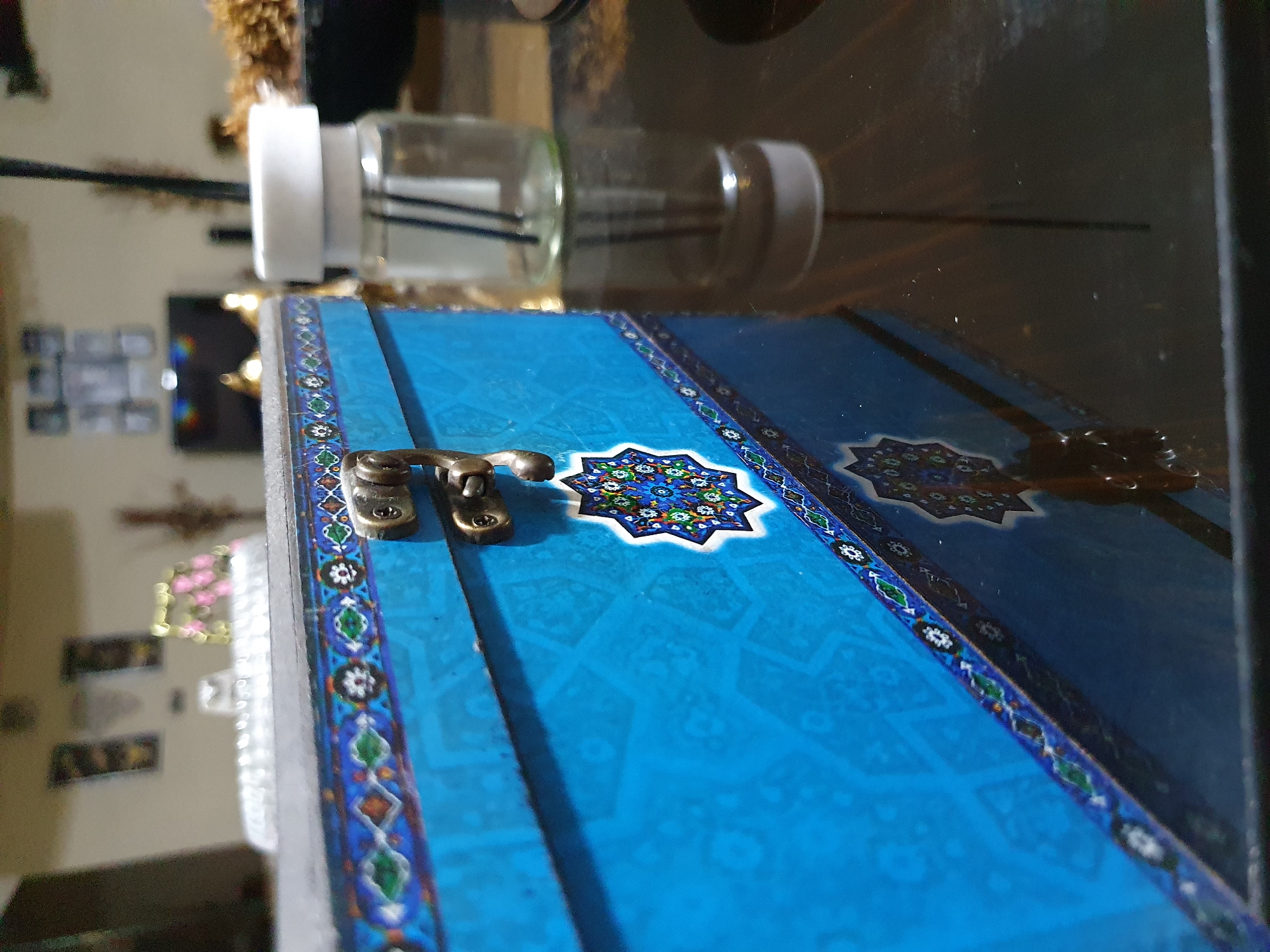 The image shows a close-up, low-angle view of a blue, decorative box with a metallic latch. The box has intricate patterns and star-shaped designs. To the right of the box, a clear glass jar with a white lid and dark reeds is visible, suggesting it might be an air freshener or diffuser. The scene appears to be indoors, possibly on a reflective surface like a table, with a blurred background that includes framed pictures on a wall and other objects, hinting at a domestic or office setting. The lighting suggests it's daytime, but artificial light sources might also be present. The overall impression is one of a still-life composition focusing on the textures and decorative elements of the box and the nearby jar. The location in Shiraz, Iran is not discernible from the visual information provided in the image.