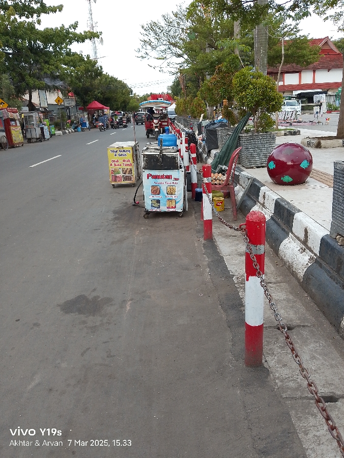 The image shows a street in Indramayu, Indonesia, located at the coordinates -6.326 latitude and 108.323 longitude. The street is paved with asphalt and lined with a sidewalk and a curb. There are trees and shrubs on the sidewalk, and a few red and white poles with a chain connected to them are lined up along the edge of the sidewalk. There are several street food vendors selling food from their carts on the sidewalk. One vendor is selling "Telur Gulung & Sosis" and another is selling "Pempek." There is a red, spherical object on the sidewalk, seemingly made of concrete. There are a few pedestrians walking along the street and some cars in the background. The street is clear and the sidewalk is relatively clean. The image is taken from the perspective of someone walking along the sidewalk.