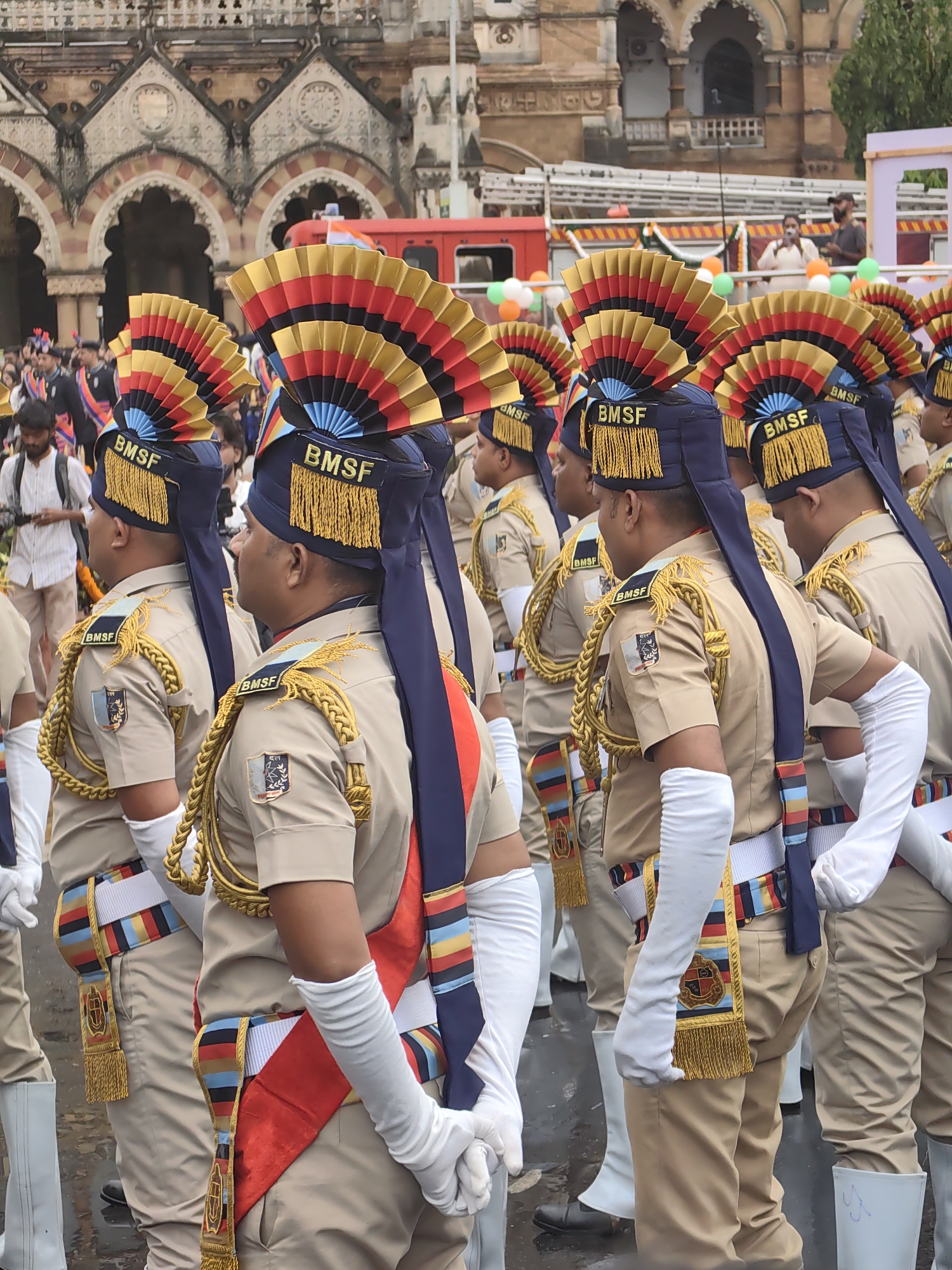 The image captures a parade in Mumbai, India, with uniformed personnel in the foreground. They are wearing tan uniforms with blue and gold embellishments, including elaborate feathered headdresses. Each uniform features a dark blue cap with the letters "BMSF" and a golden fringe. The personnel are adorned with sashes of various colors, including red, blue, yellow, and black, and some wear white gloves. A distinctive emblem is visible on their shoulders and on a sash. In the background, a historical building with arched windows and intricate architecture is partially visible, suggestive of a significant landmark. A red fire truck, adorned with Indian flags and balloons in the national colors (orange, white, and green), is also present, indicating a festive or commemorative event, likely Independence Day. The ground appears wet, suggesting recent rain. The overall scene depicts a formal procession or ceremony.
