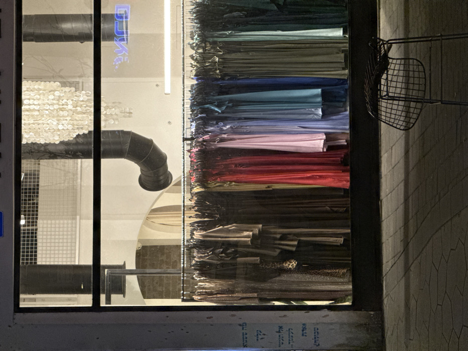 A window display showcases racks of colorful fabrics arranged by hue, from green to blue to pink to red, and finally various shades of brown. To the left, a glimpse inside the store reveals metal grates, a ventilation pipe, and hanging white discs. Outside the window, part of a metal basket and patterned pavement are visible. Text is present at the bottom of the window.