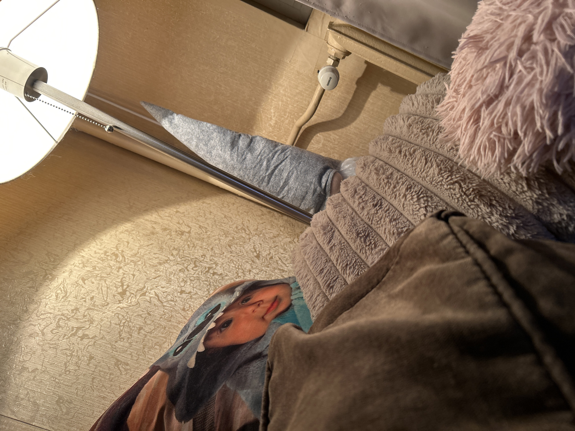 An infant is nestled amongst soft blankets, wearing a blue hooded garment with cartoonish features. A portion of a floor lamp and a radiator are visible in the background, suggesting an indoor setting. The lighting is warm and ambient. The scene is likely within a residential apartment in Sofia, Bulgaria, potentially a bedroom or living area.