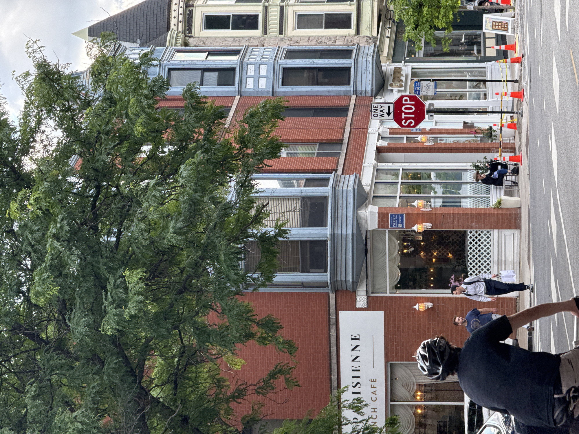 A cyclist wearing a black helmet and black shirt rides towards the viewer on a street in Chicago, Illinois. To the right, a woman sits on a bench outside a cafe, smoking a cigarette. A man walks by, looking at her. To the left, the facade of a brick building displays the sign "PARISIENNE FRENCH CAFÉ." The cafe has large windows with white curtains and decorative lighting. Across the street, a stop sign and a bus stop sign are visible. The buildings in the background feature bay windows and distinctive architectural details. A large tree with green leaves partially obscures the view of the buildings. The sky is overcast.