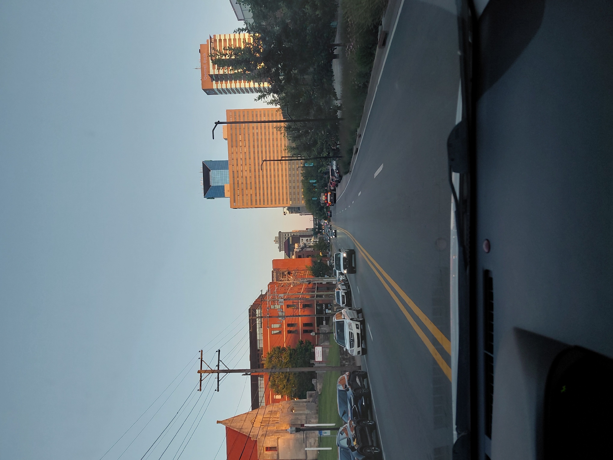 The image shows a street view in Lexington, Kentucky, USA, as seen from inside a vehicle. The perspective is from the driver's seat, looking forward through the windshield. The street is paved and has double yellow lines down the center. Several cars are driving on the road. On the left side of the street, there are parked cars, a brick building with a red roof, and a church with a sign reading "First Baptist Church." Further down the street, there are modern brick buildings and tall office buildings, including one with a sign that reads "Central Bank." The sky is clear and light blue, indicating it is either early morning or late afternoon. Trees and streetlights line the right side of the road. The overall scene depicts a busy urban environment with a mix of historical and modern architecture.