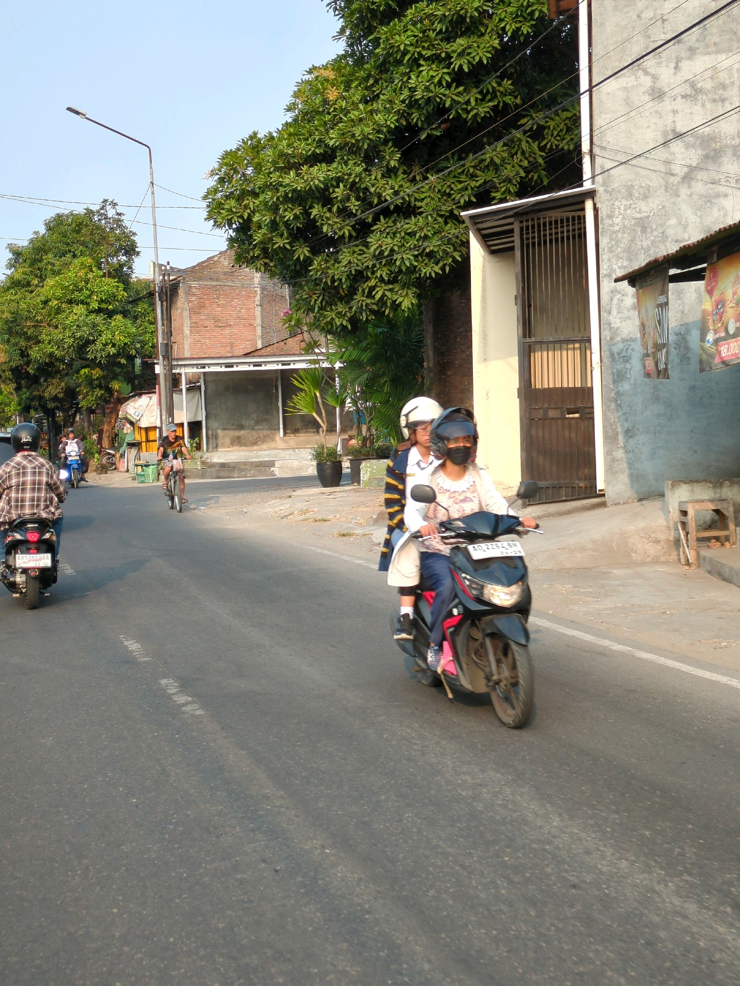 On a sunny day in Surakarta, Indonesia, two individuals are riding a black scooter on a paved road, with trees and buildings lining the street. The scooter is moving forward, with the driver wearing a white helmet and a patterned shirt, and the passenger wearing a white helmet and a striped jacket. In the background, other vehicles and people are visible. The scene depicts a typical street in Surakarta, with its blend of natural elements and urban infrastructure.