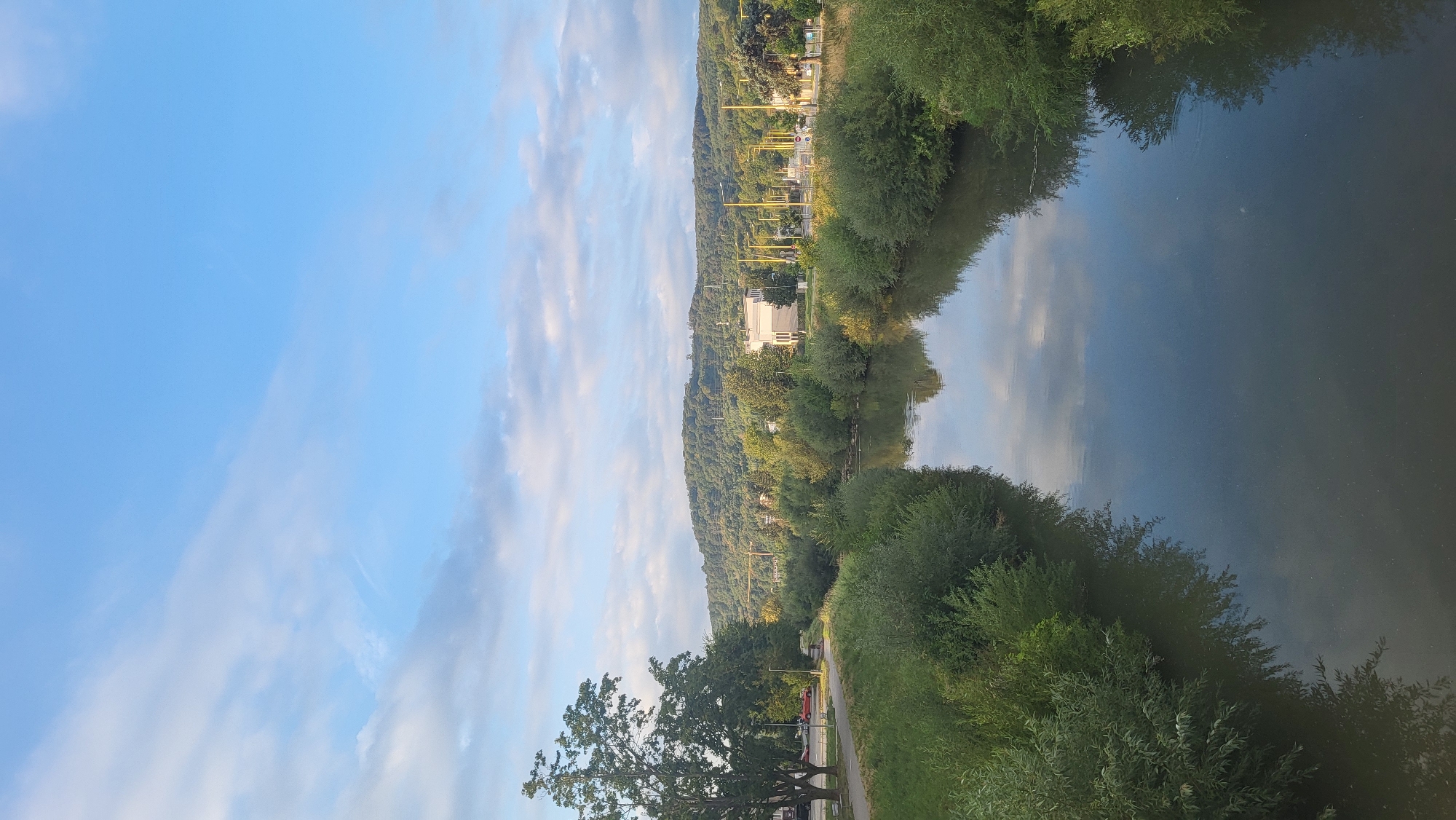 This is an outdoor, vertical, eye-level shot of a calm river flowing through a city. The river takes up the bottom half of the image, reflecting the sky and trees. On the left bank of the river is a paved pathway with a sidewalk, grass, and trees, including a large deciduous tree. Further in the distance on the left, cars are visible on a road. On the right bank, there are dense bushes and trees lining the river. Beyond the river and banks, there is a street with lampposts and some buildings, including a white rectangular building. In the background, a tree-covered hill rises under a bright blue sky with scattered white clouds. The image appears to have been taken during the day. This scene is the Torysa River in Prešov, Slovakia, as evidenced by the view of the hills and urban features consistent with the area.