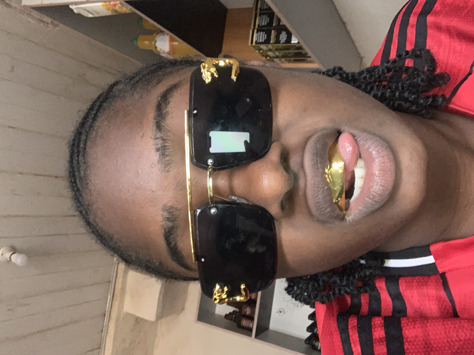 A close-up, portrait-oriented shot, rotated to appear vertical, features an individual with dark skin, positioned to the right of the frame. The person is wearing large, black rectangular sunglasses with intricate gold frames and decorative golden elements on the top outer corners, possibly depicting small animals or ornate designs. One upper front tooth is adorned with a prominent gold grill, visible through slightly parted lips. The individual's hair appears to be styled in braids or cornrows. They are wearing a red garment with black vertical stripes, characteristic of athletic wear.
The background suggests an indoor setting, likely a shop or concession area near the New Stadium in Nigeria. To the upper left and behind the person's head, light-colored wooden shelves are visible, holding bottles with yellow liquid and other packaged goods. A small white lightbulb is affixed to the wall on the far left. To the upper right, another darker shelving unit with various items can be seen. The overall lighting is consistent with an indoor environment. The image captures the individual posing, highlighting their distinctive style and accessories. No discernible text is visible in the image.