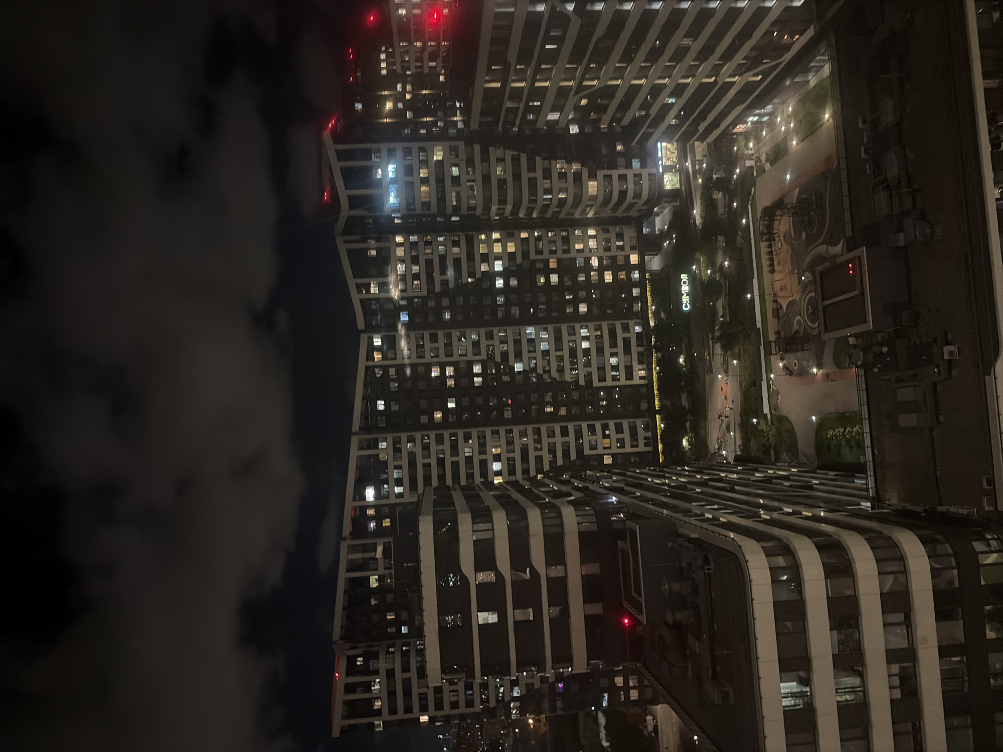 This image depicts a nighttime aerial view of the residential complex "Symvol" located in Moscow, Russia, specifically in the Basmanny District. The complex consists of multiple modern, multi-story buildings with distinctive architectural designs, characterized by curved facades and numerous illuminated windows. The buildings are arranged around a central courtyard, which features a playground, pathways, and green spaces, all illuminated by artificial lighting. Several tall buildings are visible in the background, suggesting an urban environment. The sky is dark and partly cloudy, with some red lights visible atop some of the buildings, likely aircraft warning lights. The overall scene conveys a sense of urban development and nighttime activity within a residential area.