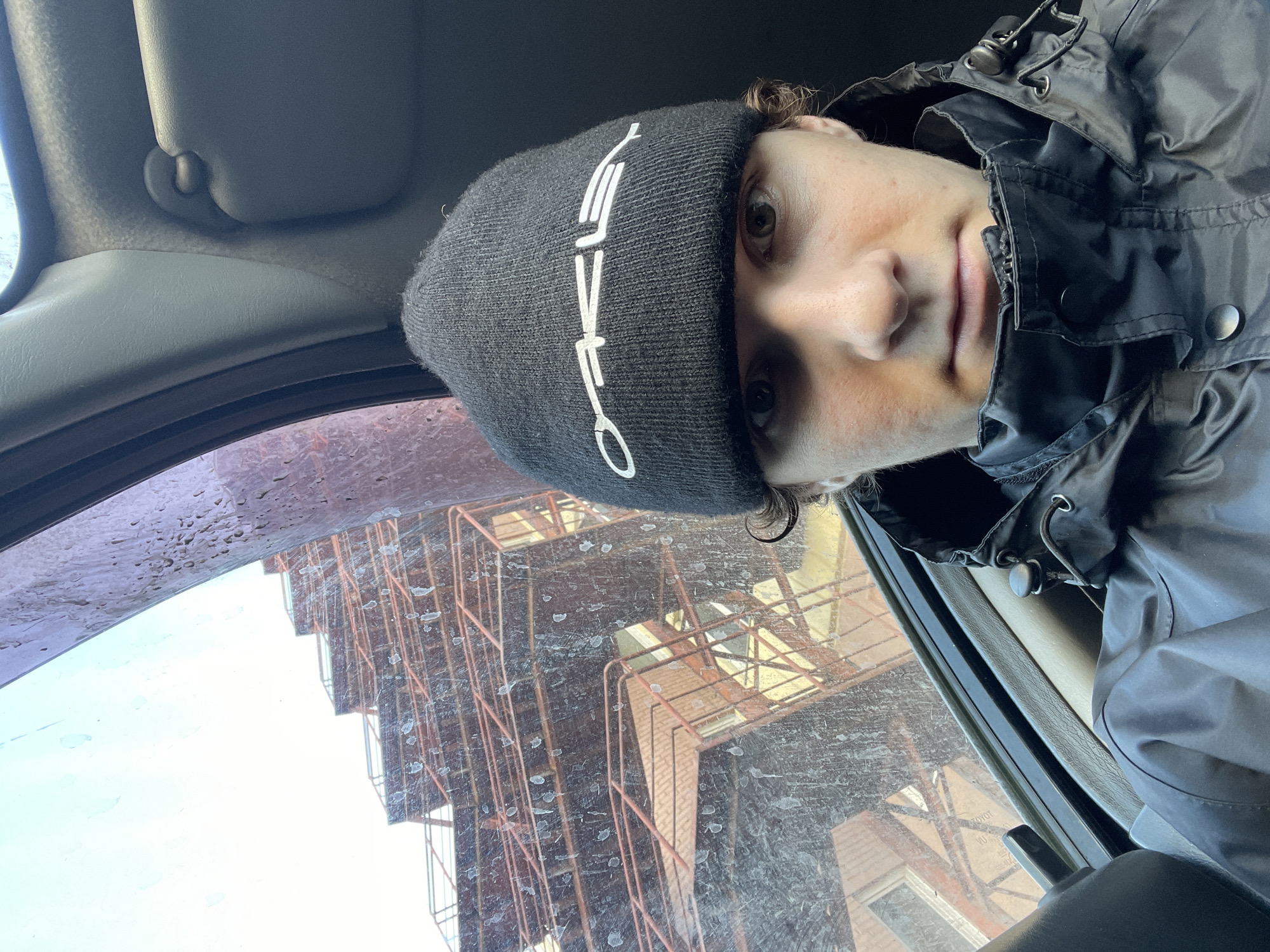A selfie taken from inside a vehicle in Krasnoyarsk, Russia. The individual is wearing a black Oakley beanie and a black hooded jacket. The vehicle's interior is visible, including the sun visor. The windshield is dirty, and the reflection shows scaffolding and a brick building. The image captures a moment in the city, possibly near a construction site.